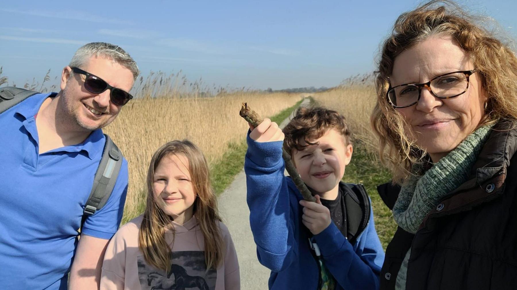 Family photo, two adults and two children out for a walk on a coastal path.  Blue sky and wheat field in the background. 