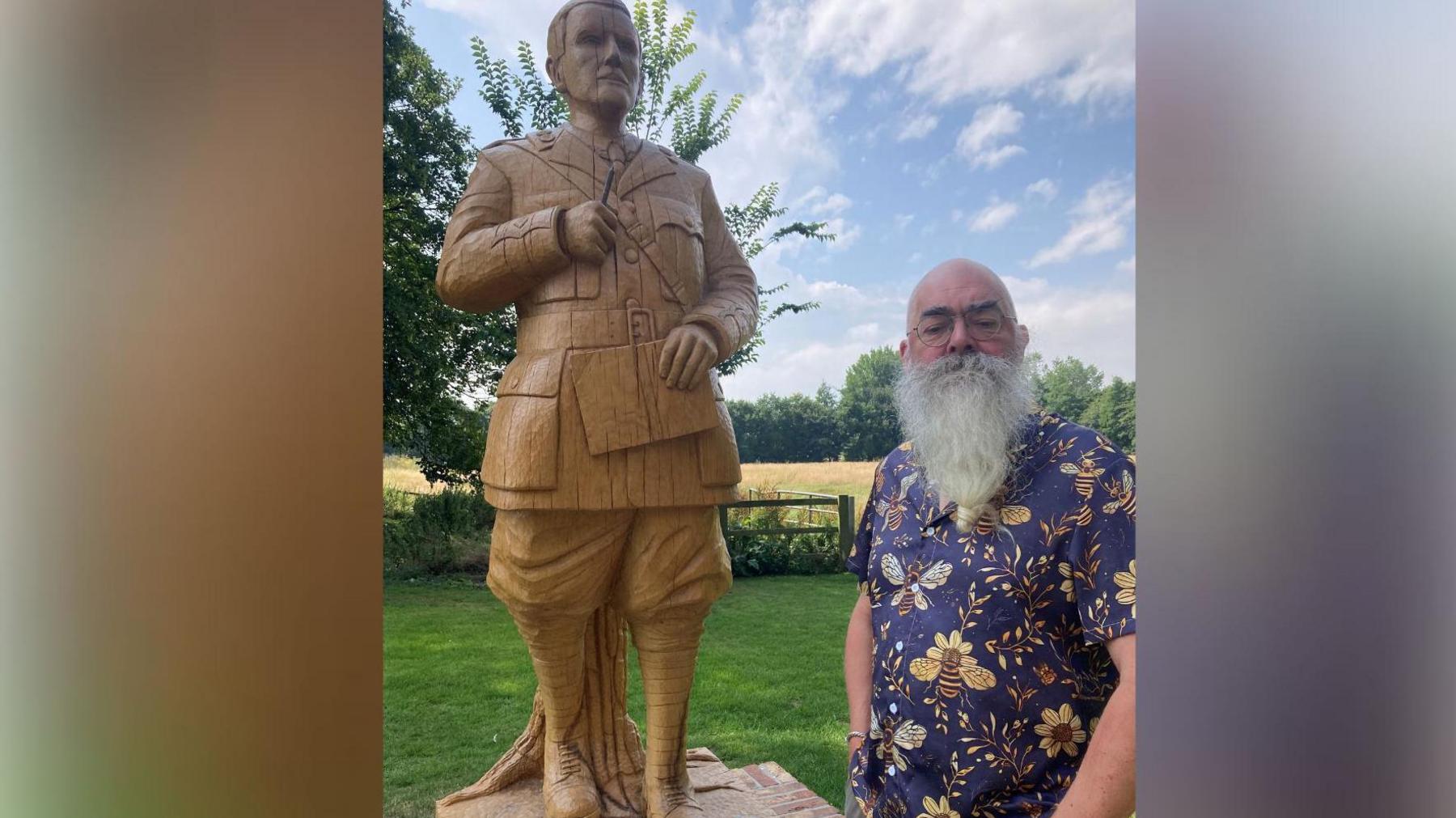 A bearded man in a blue shirt decorated with yellow bees stands next to a wooden statue of JRR Tolkien, that is showing cracks.