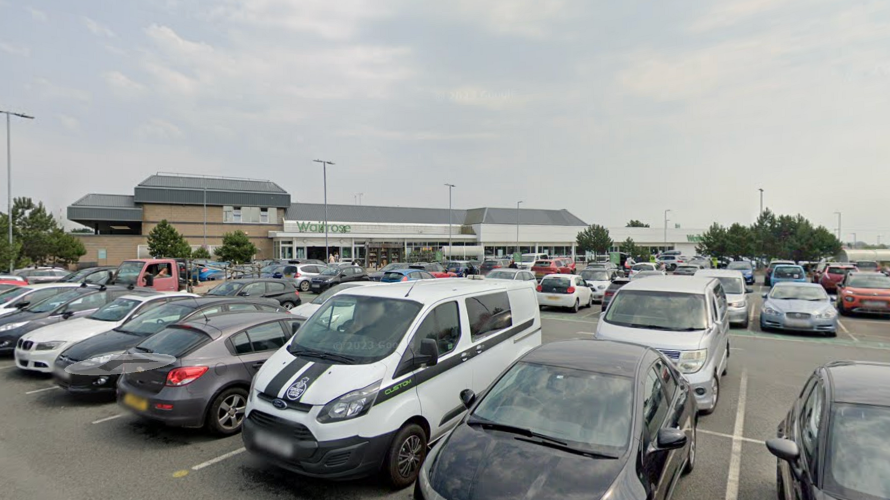 A google street view image of a Waitrose car park. There are cars and vans parked in the car park. In the background is a Waitrose building. There are trees and lampposts in the car park.