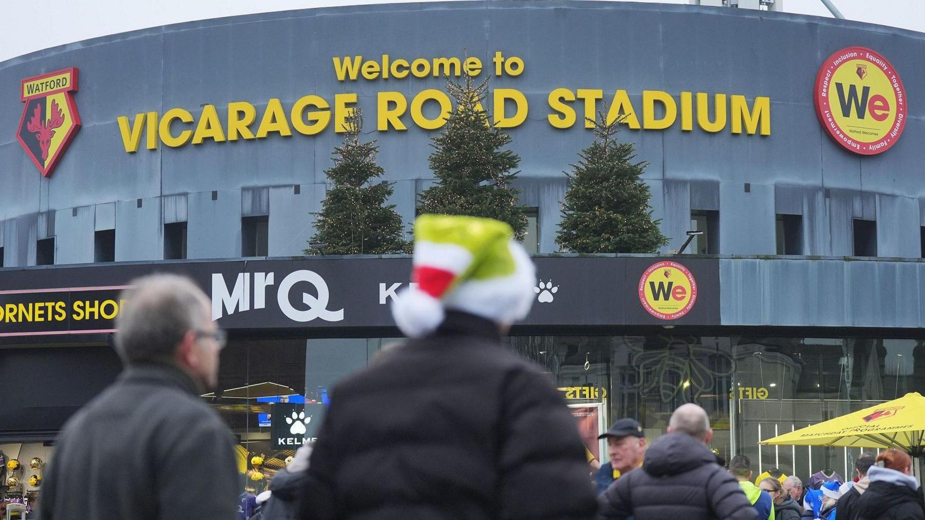 Vicarage Road stadium with its name in yellow lettering and the Watford logo on the left (a red stag with large horns, against a black and yellow background). In the foreground of the grey-coloured stadium are fans seen from behind, including one wearing a green and red Santa hat.