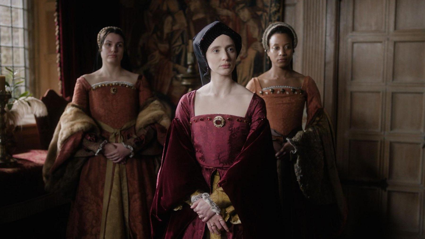 Three women in ornate, regal dresses look serious with their arms folded in front of them. 