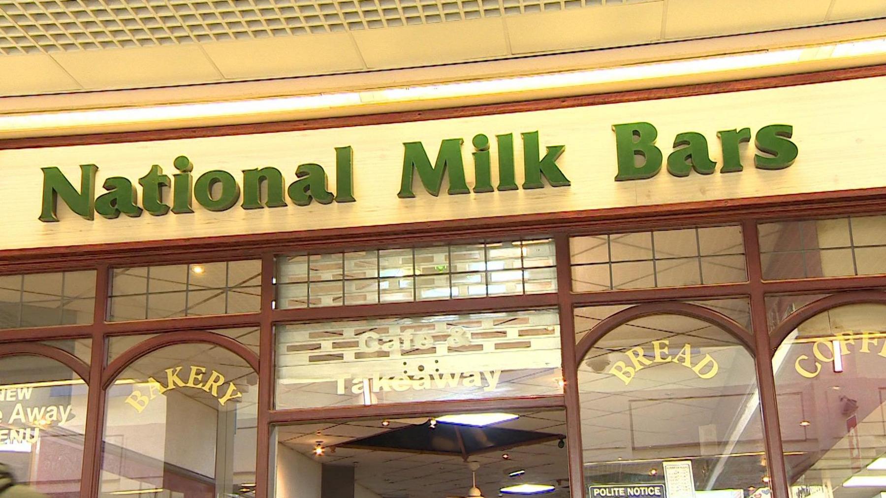 A cafe sign in green which reads ' National Milk Bars' with a glass frontage with arched windows