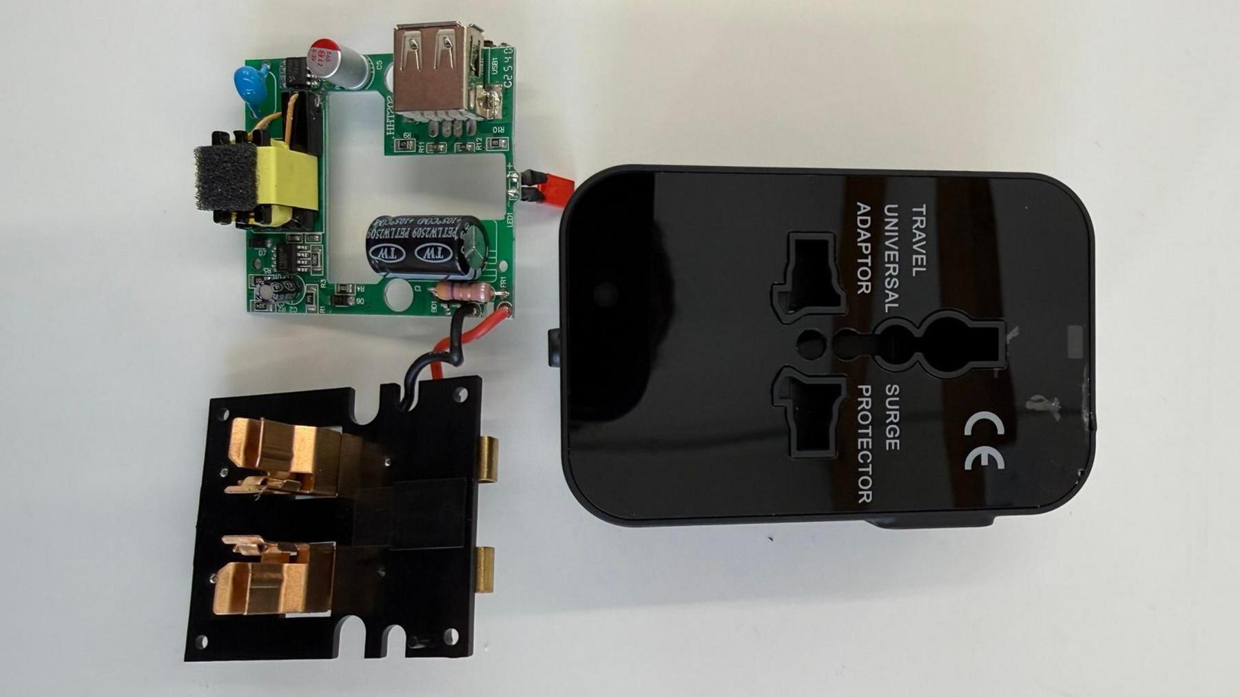 A circuit board and small plastic cover on a table beside a black, plastic travel adaptor. Text on the adaptor case, next to its sockets, reads "TRAVEL UNIVERSAL ADAPTOR" and on the other side: "SURGE PROTECTOR". A "CE" certification standard symbol is also displayed on the casing.
Copyright