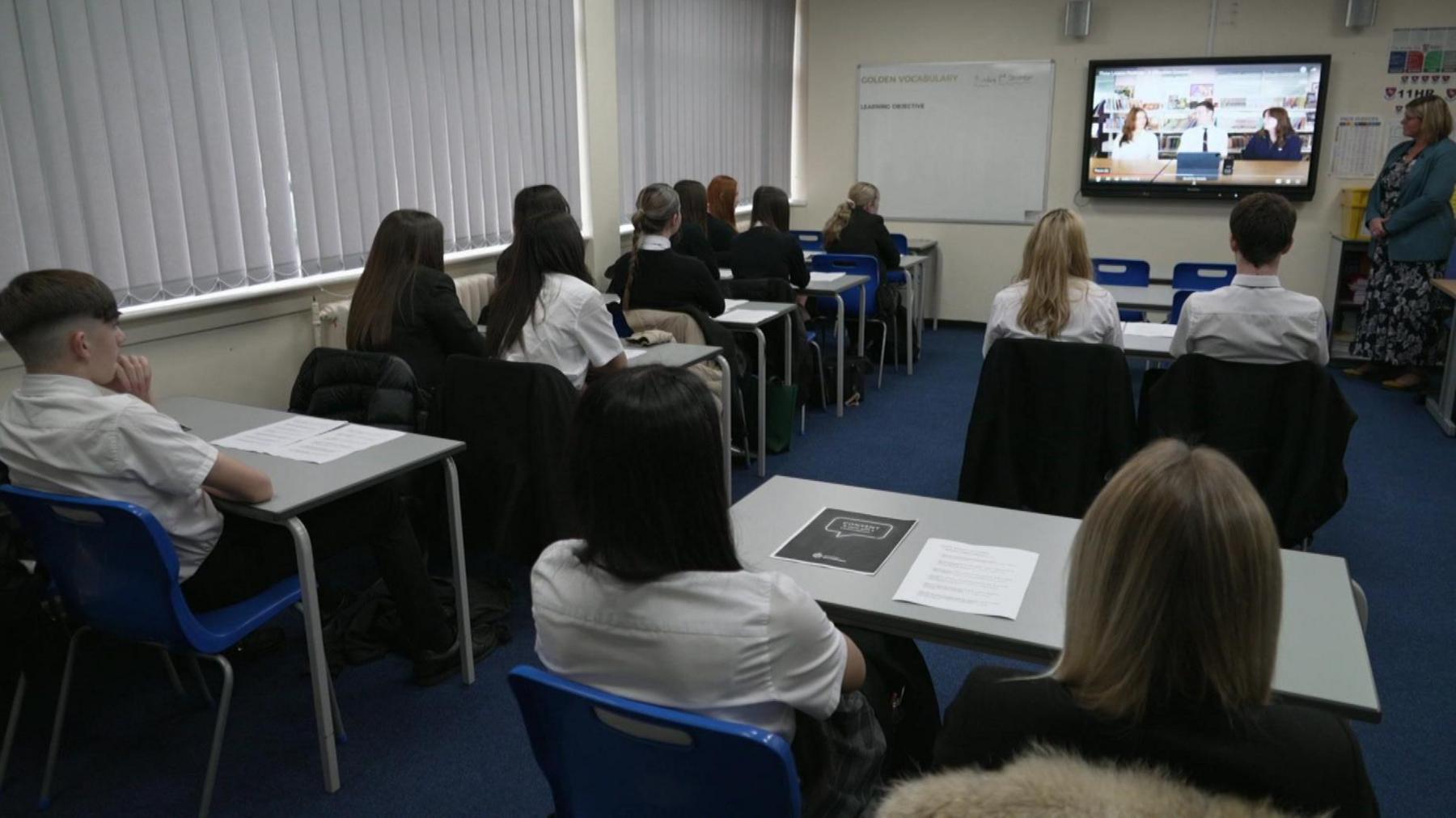 Classroom of students sat at desks watching a video