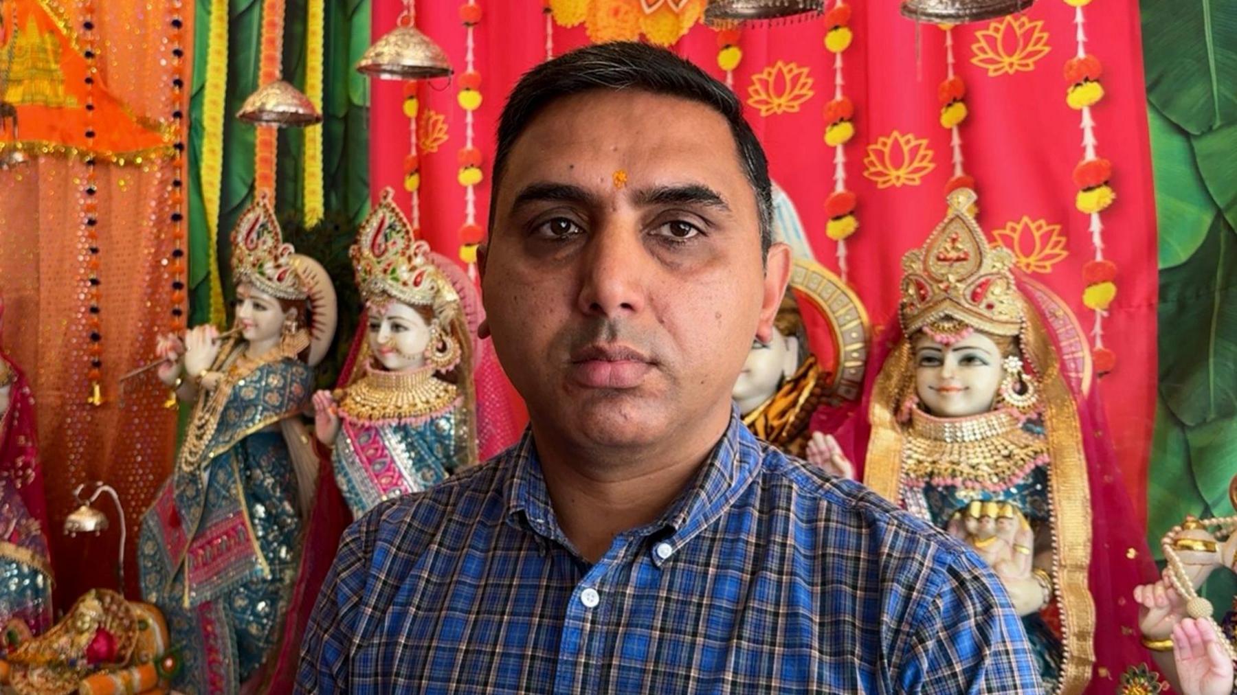 A man wearing a blue checked shirt. He is stood in front of brightly coloured wall hangings and small statues of people with Hindu clothing on.
