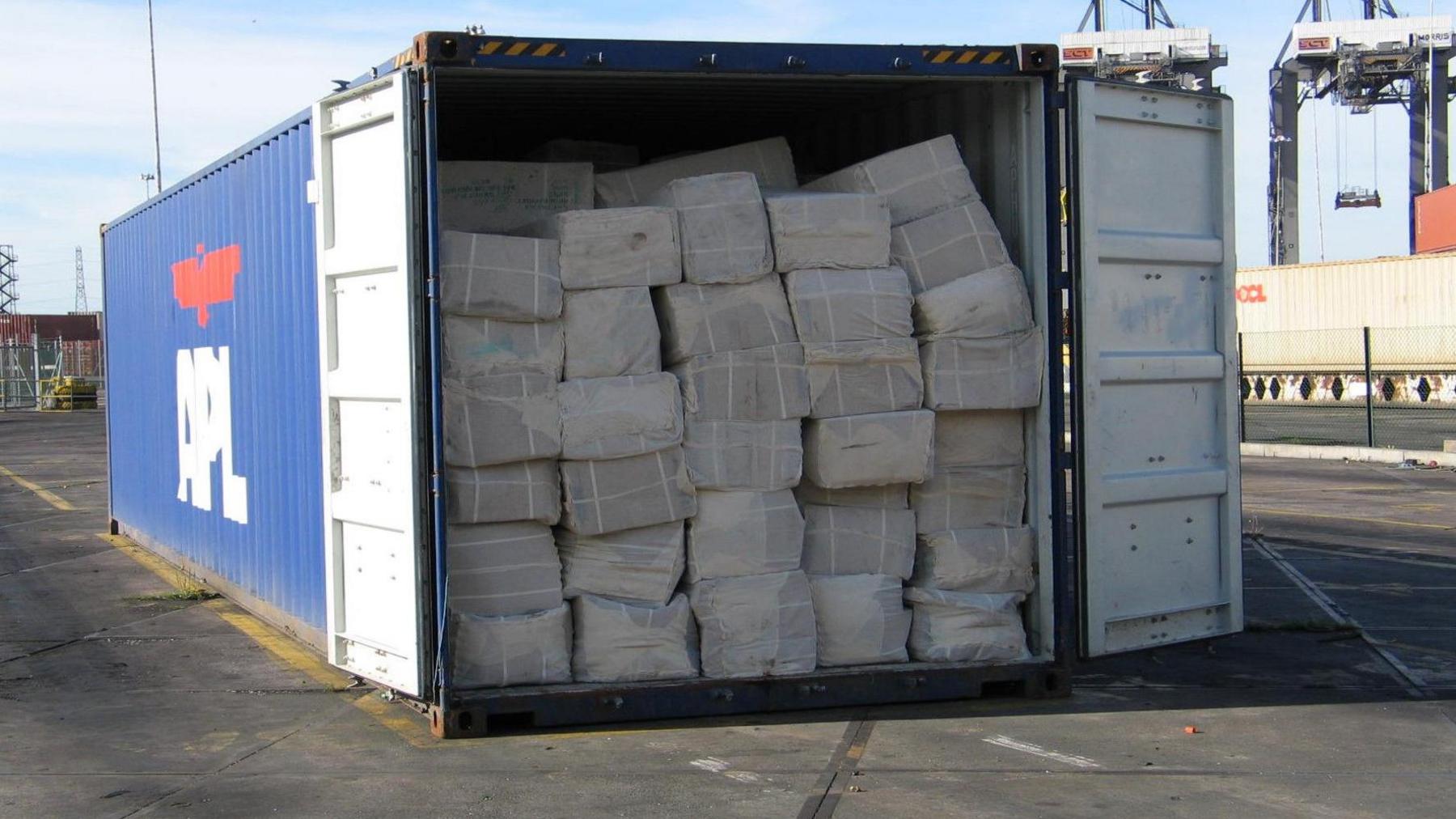 A container ship container packed full of bundles of counterfeit clothes sits on a dockside, with its door wide open