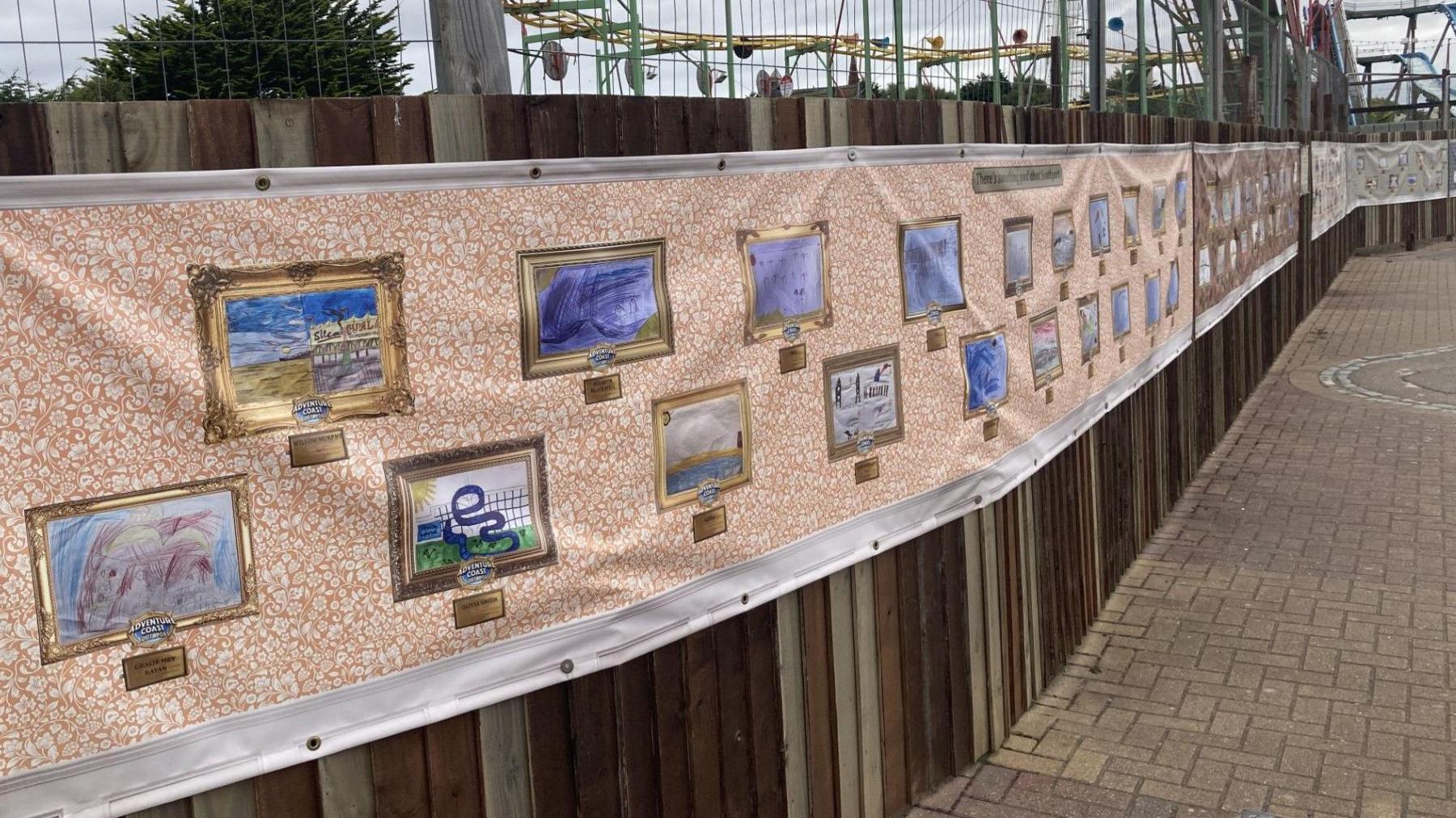 A landscape photo of some of the artwork on display. The pieces have been printed on to a long plastic sheet which is pinned to a wall at the amusement park. 