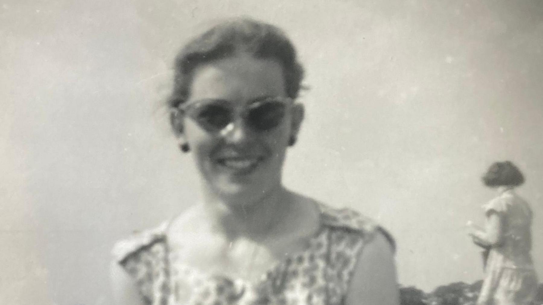 A black and white photo of a young woman wearing sunglasses and a summer dress, with another woman in the background facing away from the camera.