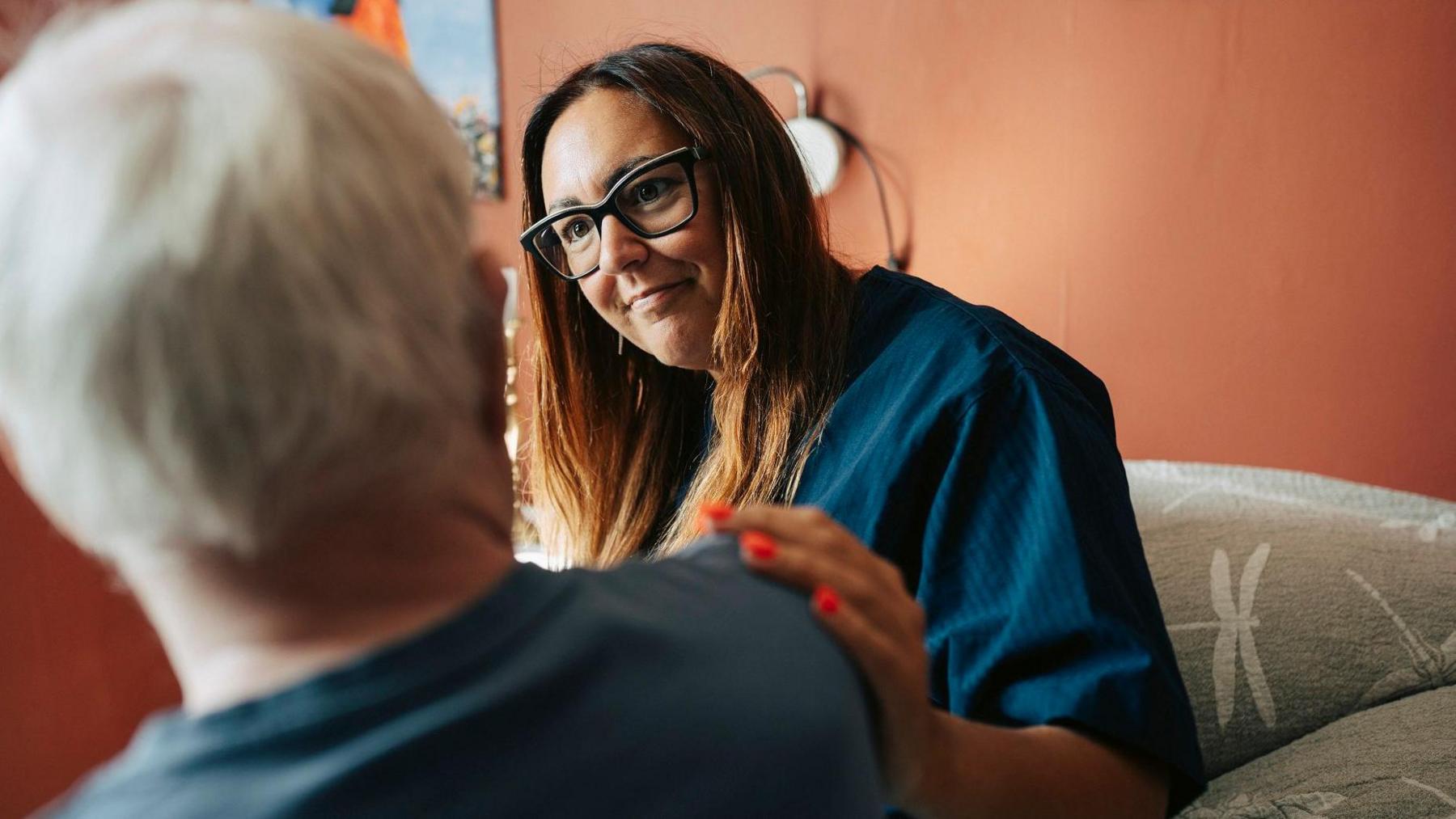 A woman with long brown hair, wearing glasses and a blue shirt, smiles as she places a hand on the shoulder of a man with short white hair, who has his back to the camera. They are sitting on a grey sofa in a lounge with copper-coloured walls.