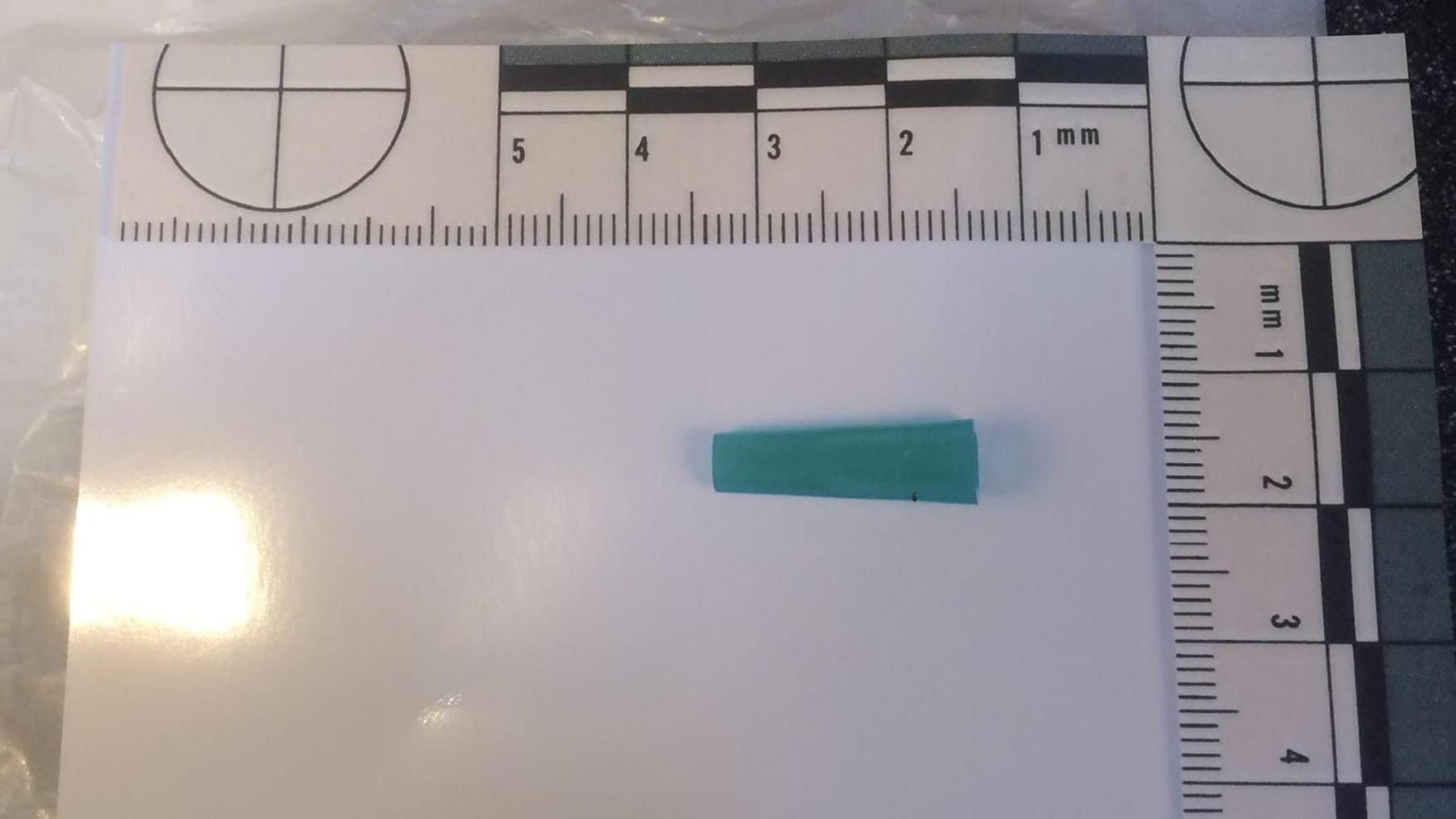 A green needle cap lies on a piece of paper. It measures about 2cm long. There are ruler lines and measurements on the X and Y axis.