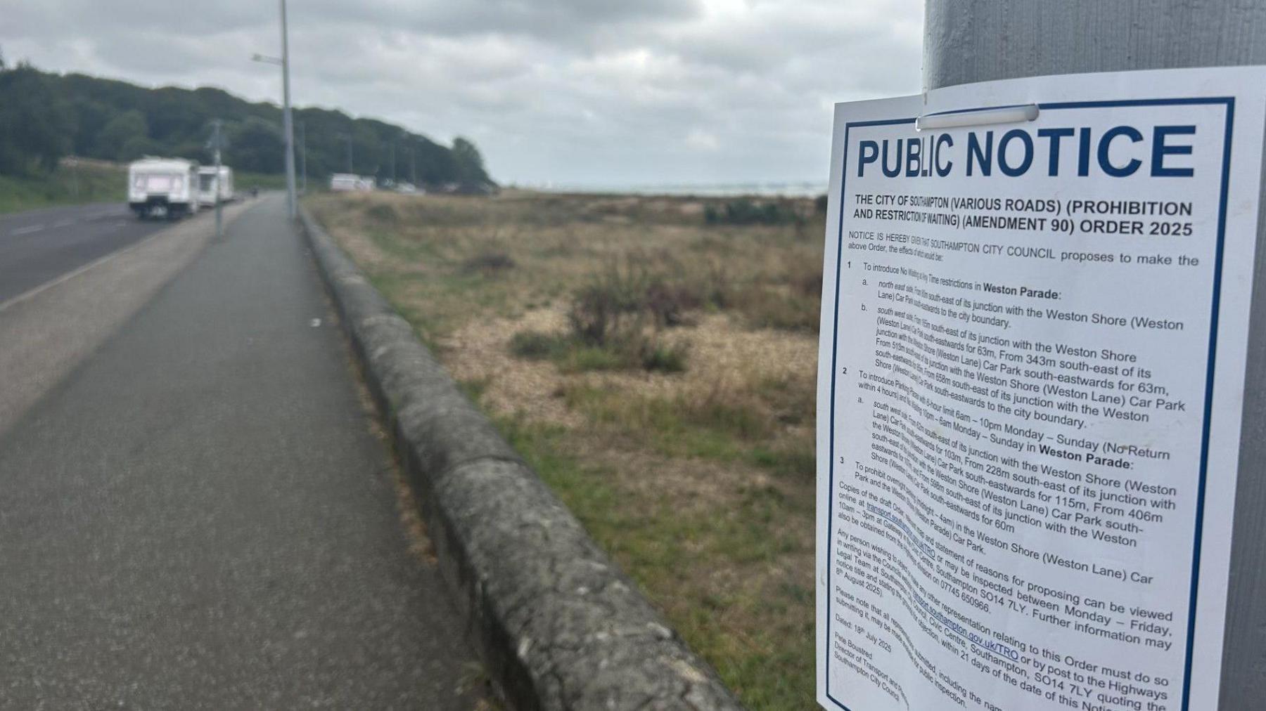 Public notice sign on the right attached to a lamppost with path and road where several caravans are parked in the distance.