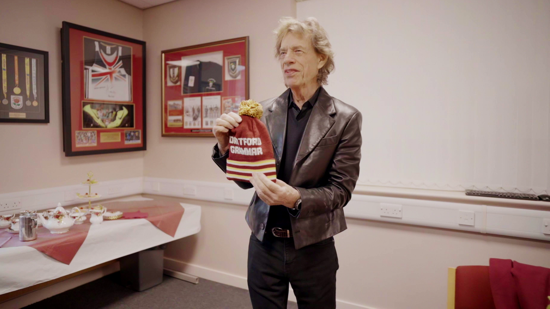 Sir Mick Jagger wearing a dark shirt and leather jacket, holding a burgundy bobble hat with a yellow bobble and white stripes at the bottom. Text on the hat reads Dartford Grammar. Framed sports memorabilia is on the wall behind him and there is a china tea set laid out on a table with white and burgundy tablecloths.