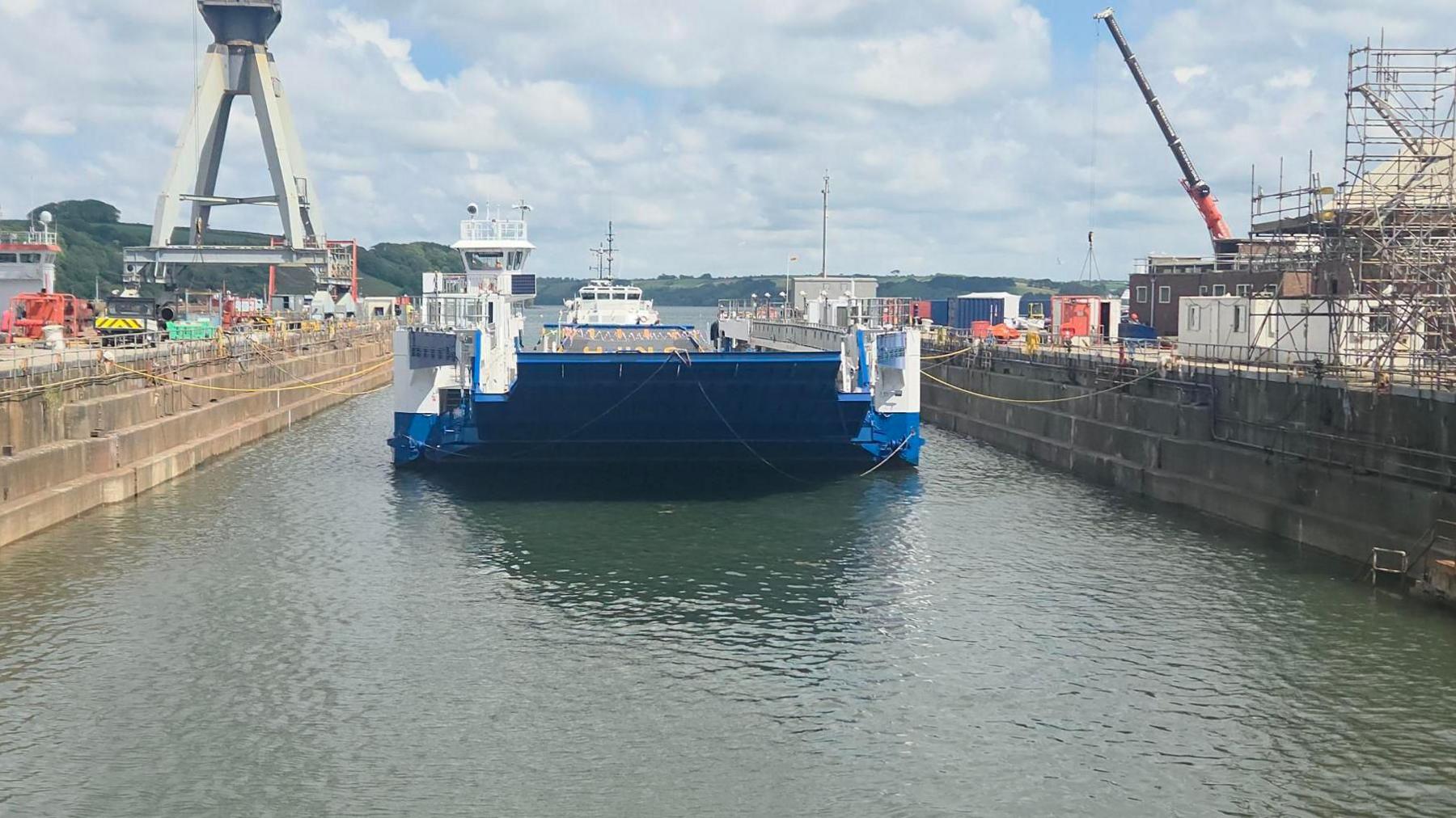 The vessel Lynher in the dry dock, except the dry dock is filled with water. The boat is blue and white and is tied to the sides of the dock. There is scaffolding on the dock on the right-hand side and a crane. The water is a murky blue. There are clouds in the sky.