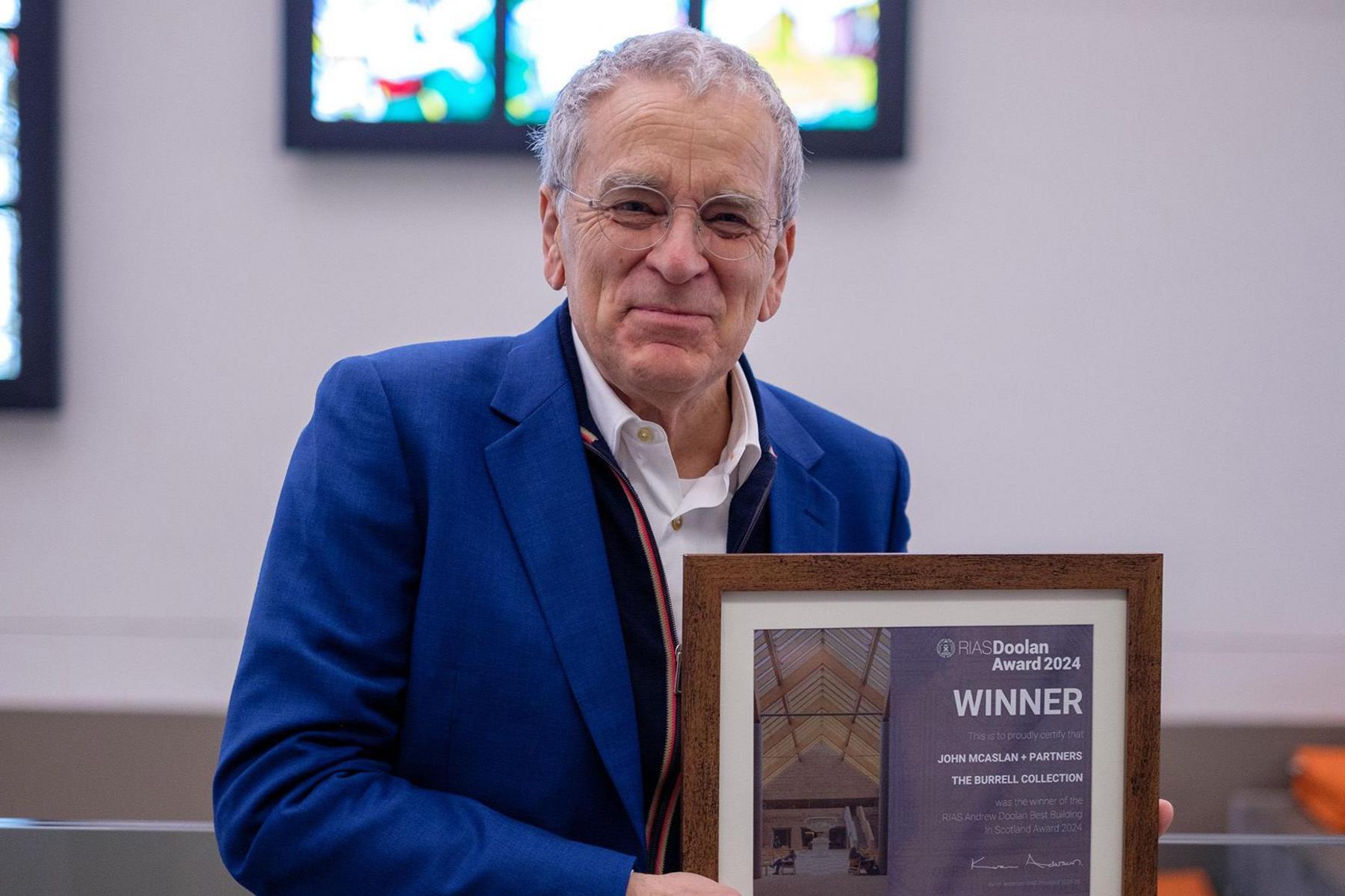 John McAslan in a blue jacket stands in front of the Burrell's stained glass holding the Andrew Doolan award - a framed plaque with "winner" printed on it.