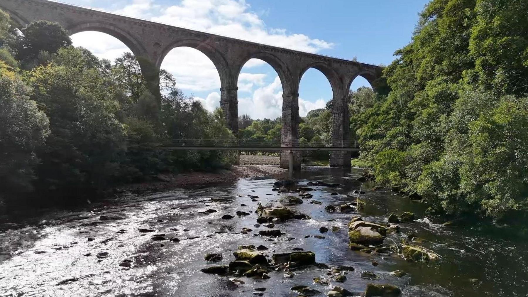 A viaduct with five arches spanning a wide but shallow and rocky river. On either side trees reach down to the water, under a blue and cloudy sky.