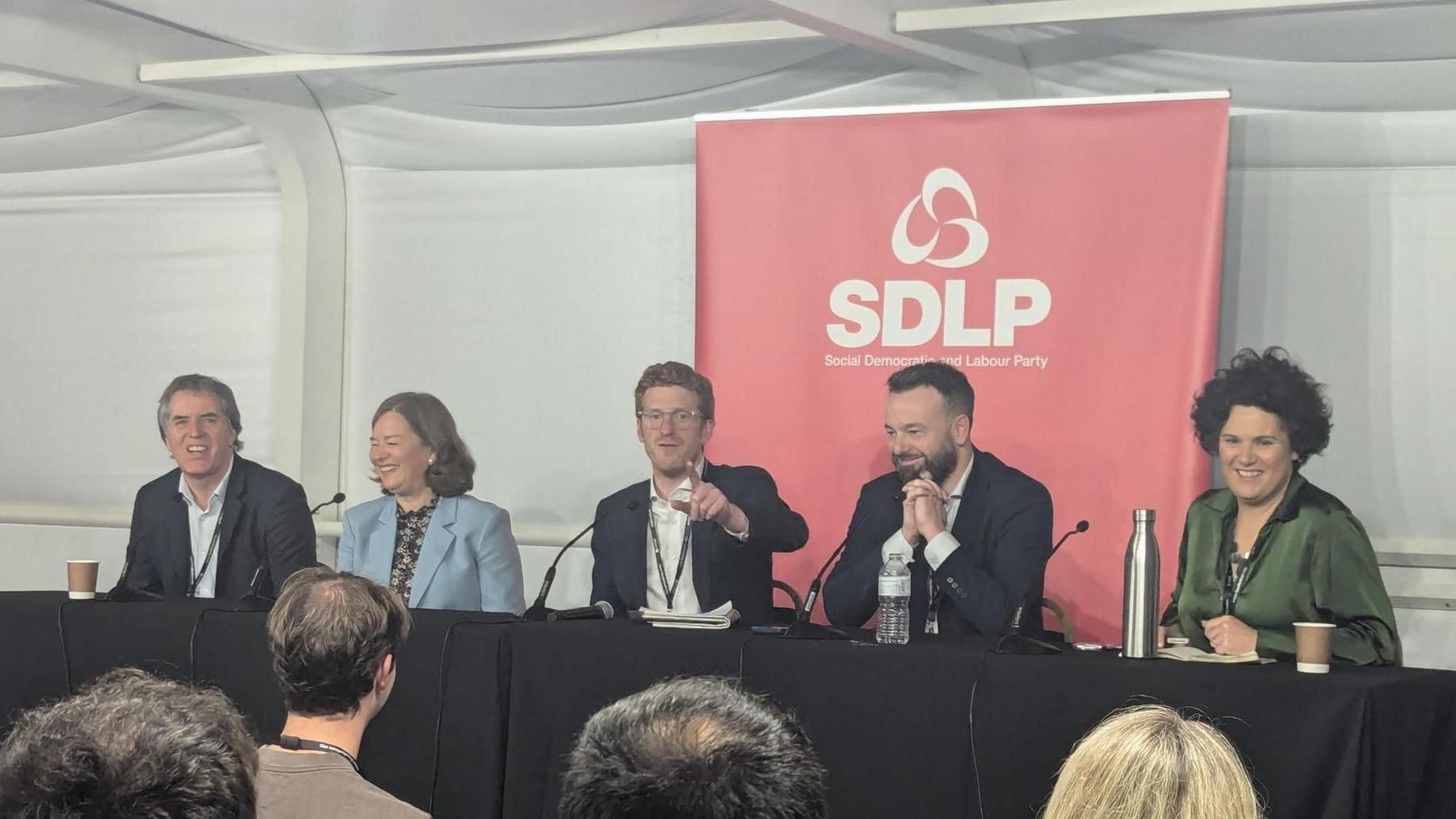 Five people sitting at a long table second in from the left is Fleur Anderson who is wearing a blue blazer with a printed top, Matthew O'Toole is dressed in a navy suit with a white shirt, Colum Eastwood wears a similar suit and Claire Hanna is dressed in green.