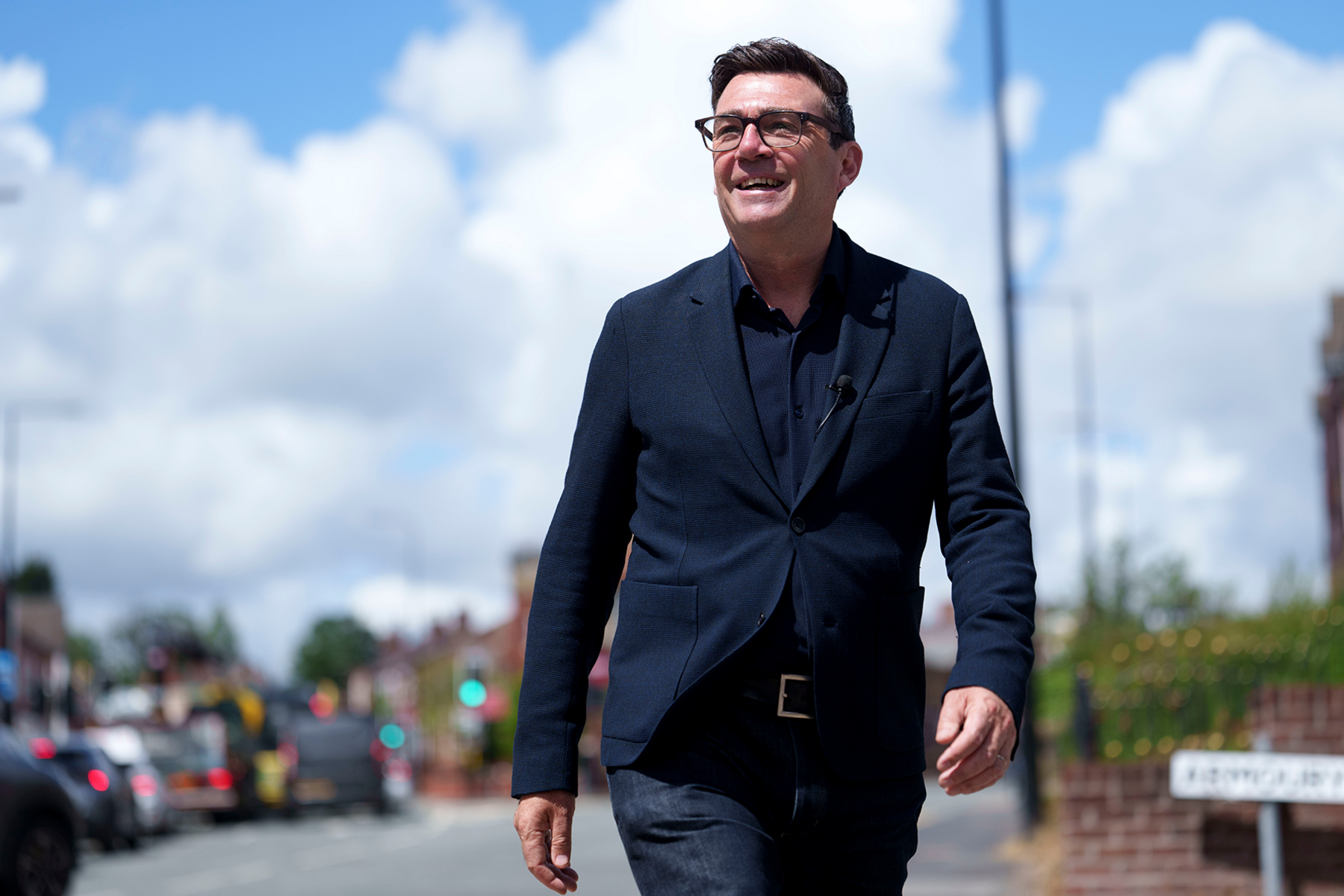 Andy Burnham smiles during a campaign visit to Ashton-in-Makerfield before the forthcoming by-election, in Manchester, England, Tuesday, June 9, 2026. He is wearing a dark suit jacket with a dark shirt underneath. He is smiling and waling in a sunny street. His hair is dark and he is wearing dark-framed glasses.