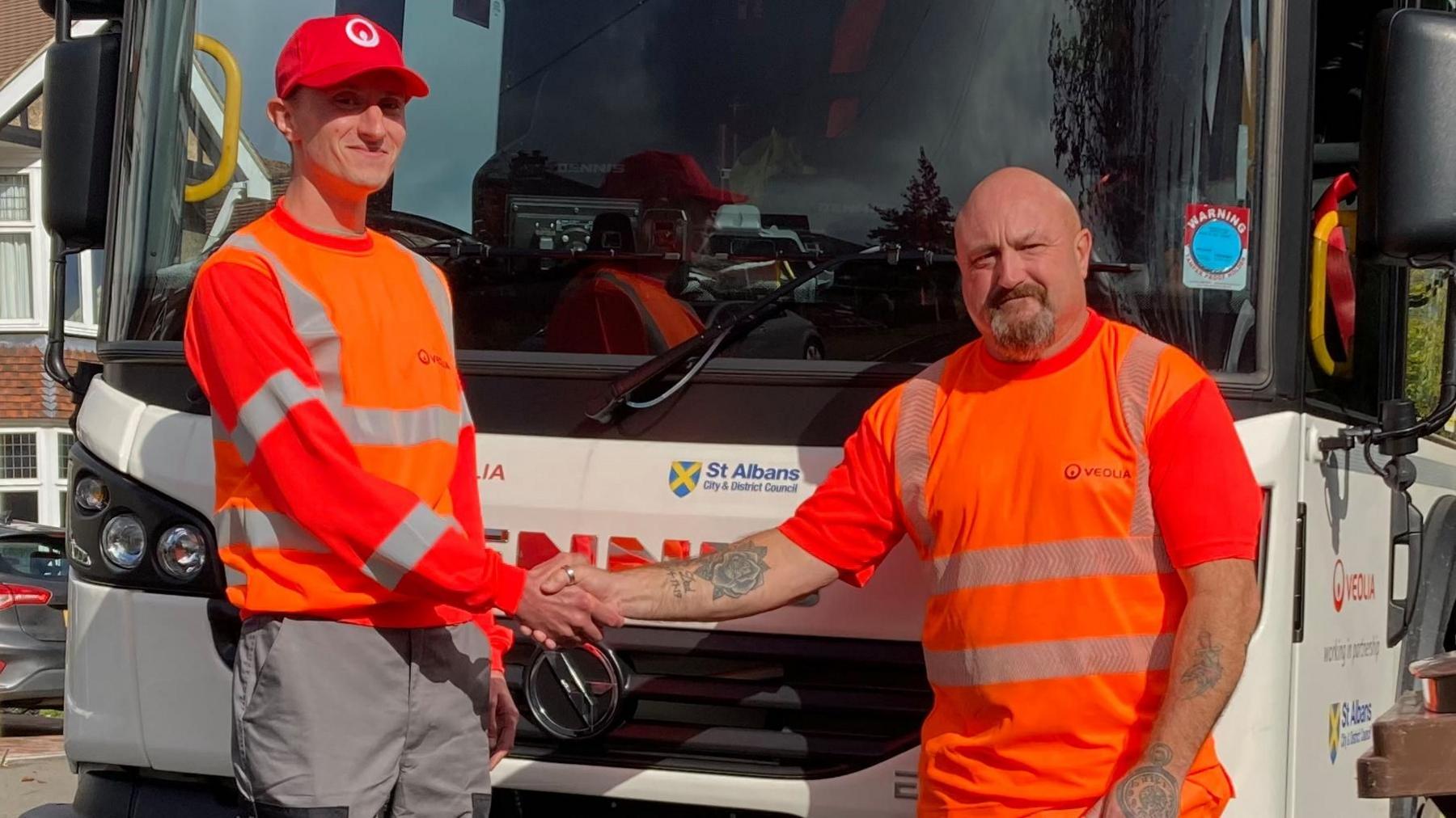 Two refuse collectors dressed in orange are stood next to a white St Albans bin lorry. they are shaking hands.