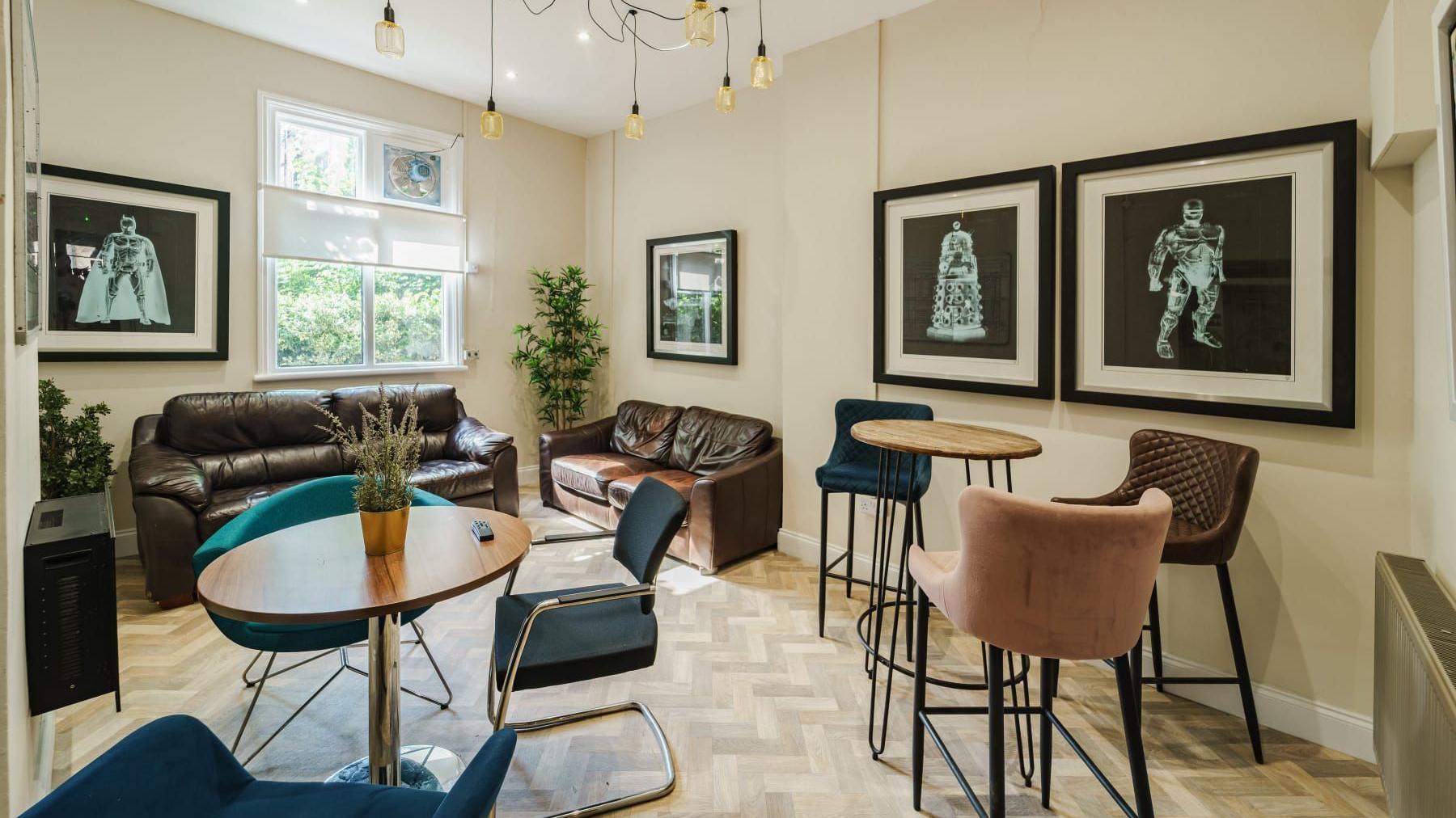 A room with two brown sofas against the walls, then a table with teal chairs and a higher table with three bar stools. The room has pale herringbone flooring and cream walls.