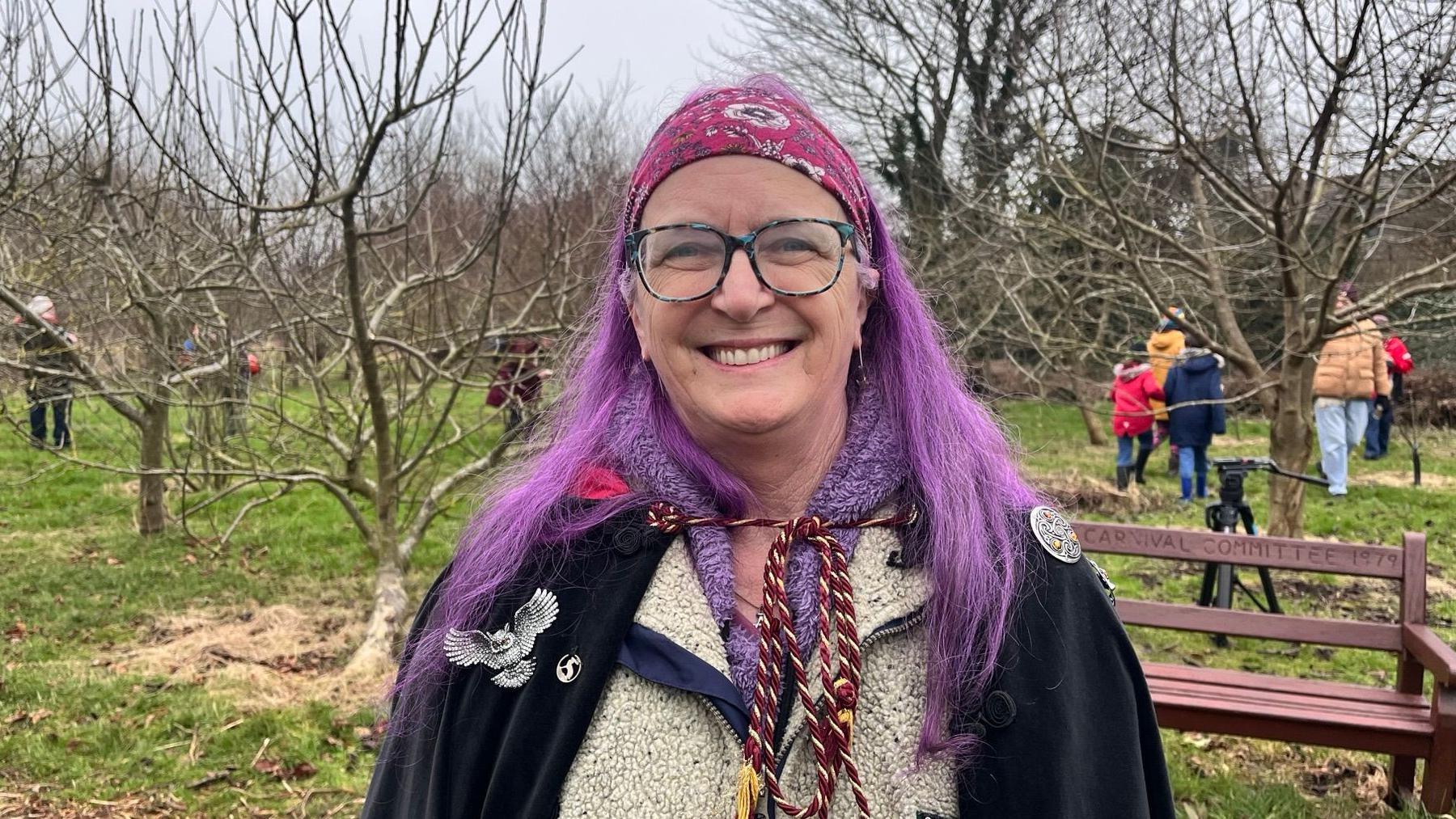 Ms Rawdon wears a black cloak with an owl on it.  She has a purple jumper underneath and bright-purple, long hair, which is kept off her face with a pink hairband.  She wears glasses and is smiling.