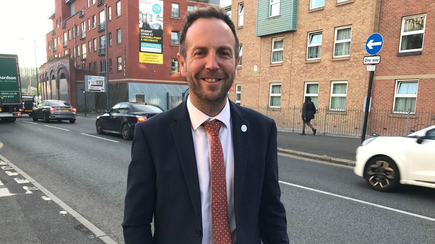 Mr Miskell stands next to a busy road, wearing a dark suit and a red tie with white dots. He is smiling at the camera, his eyes creasing at the corners. He has blond hair and sports a stubble