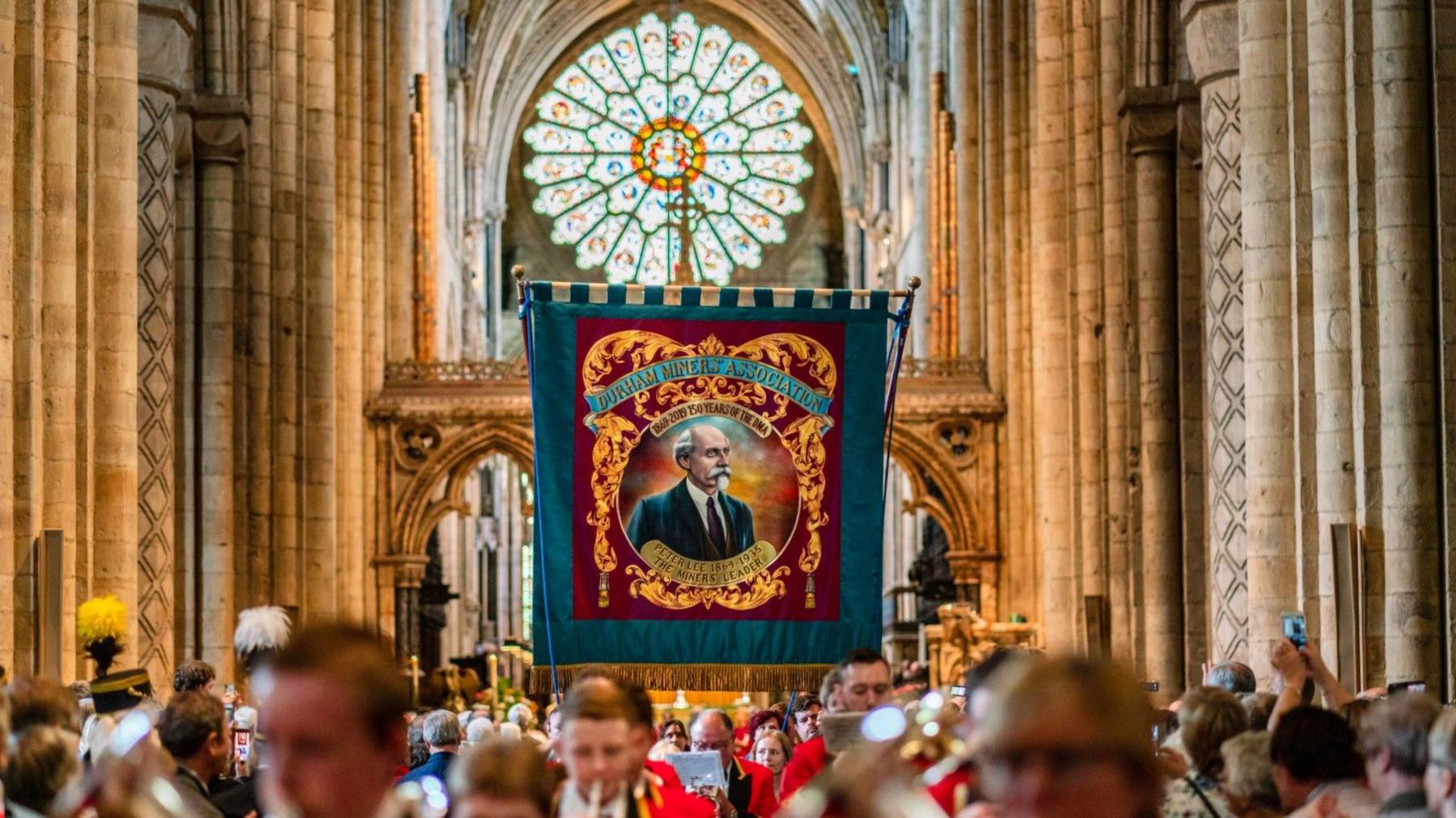 A blue miners' banner is held up in the middle of Durham Cathedral, while a brass band dressed in red clothing play music. Onlookers are taking pictures of the banner.