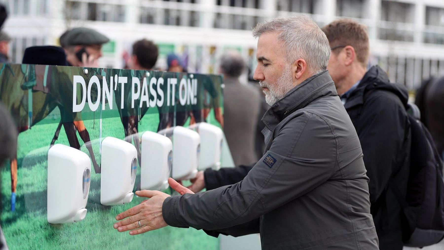 Two men at a row of hand sanitisers at Cheltenham Racecourse on 10 March 2020. The board behind the sanitisers says "Don't pass it on".