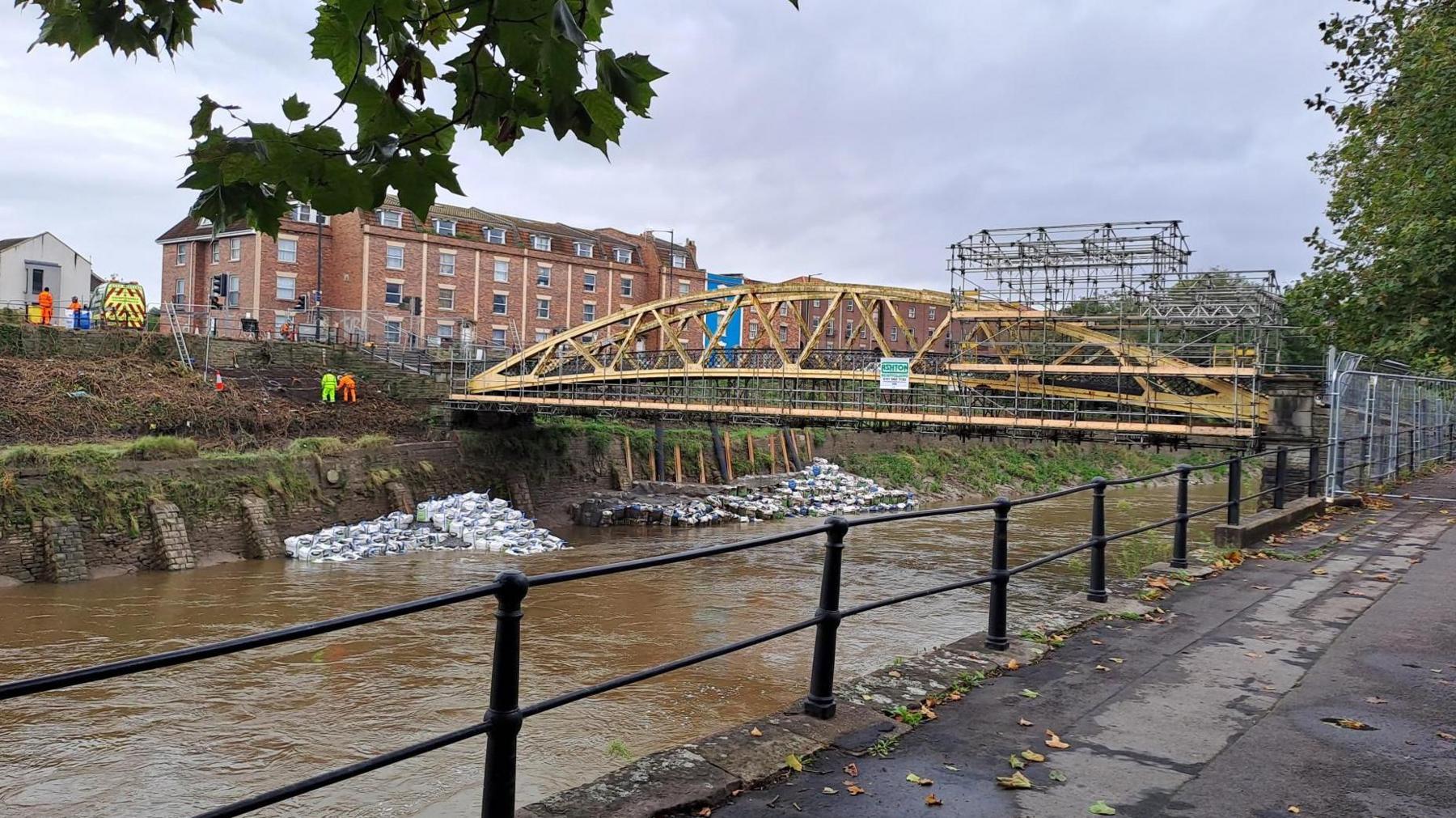 Hundreds of white sandbags piled up against the riverbank beneath Langton Street Bridge, which is a yellow footbridge across the river shaped like a banana. 