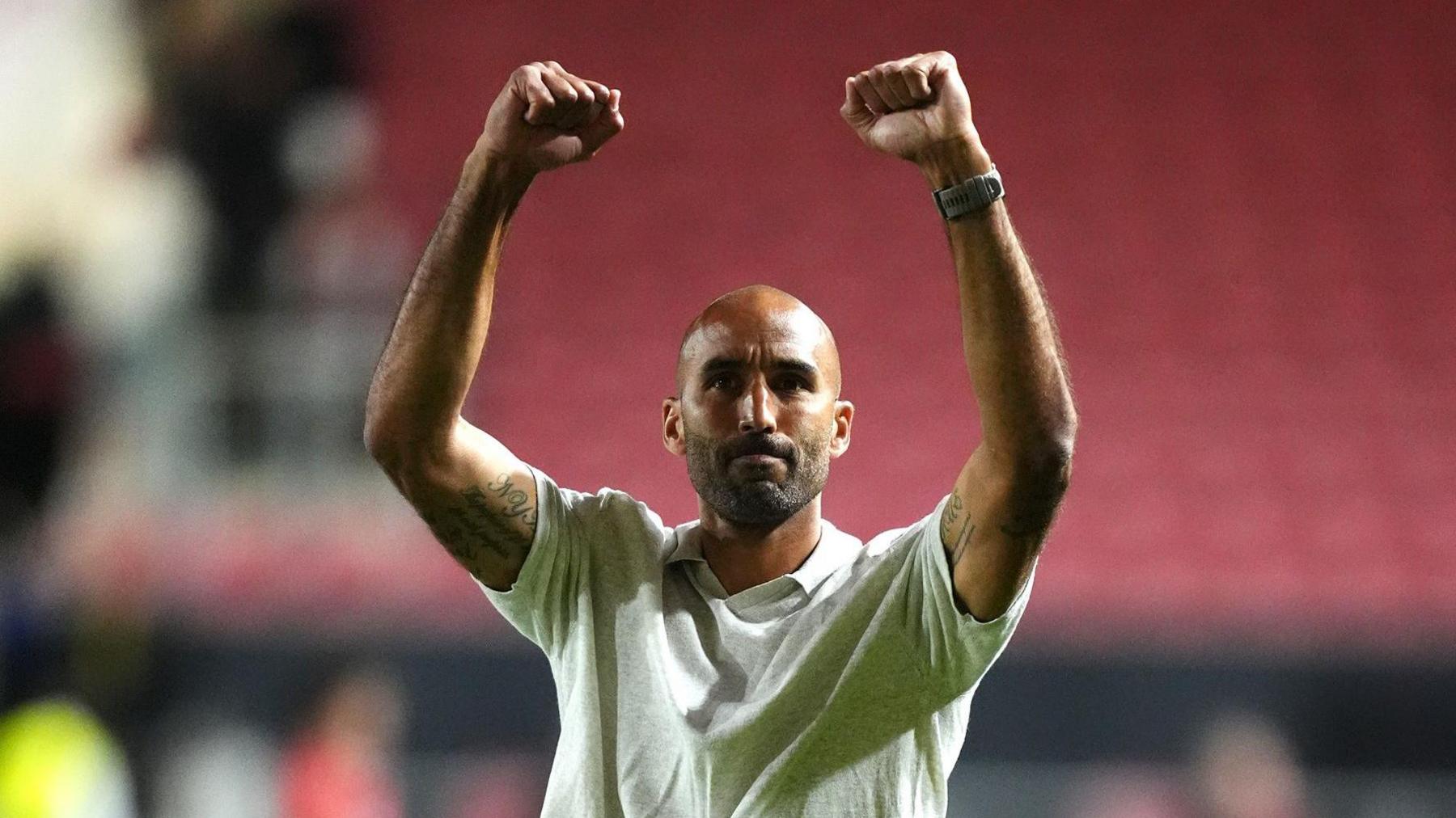 Walsall head coach Lee Grant raises both arms in celebration to the club's fans after their EFL Cup win over Bristol City. He is wearing a cream coloured t-shirt.