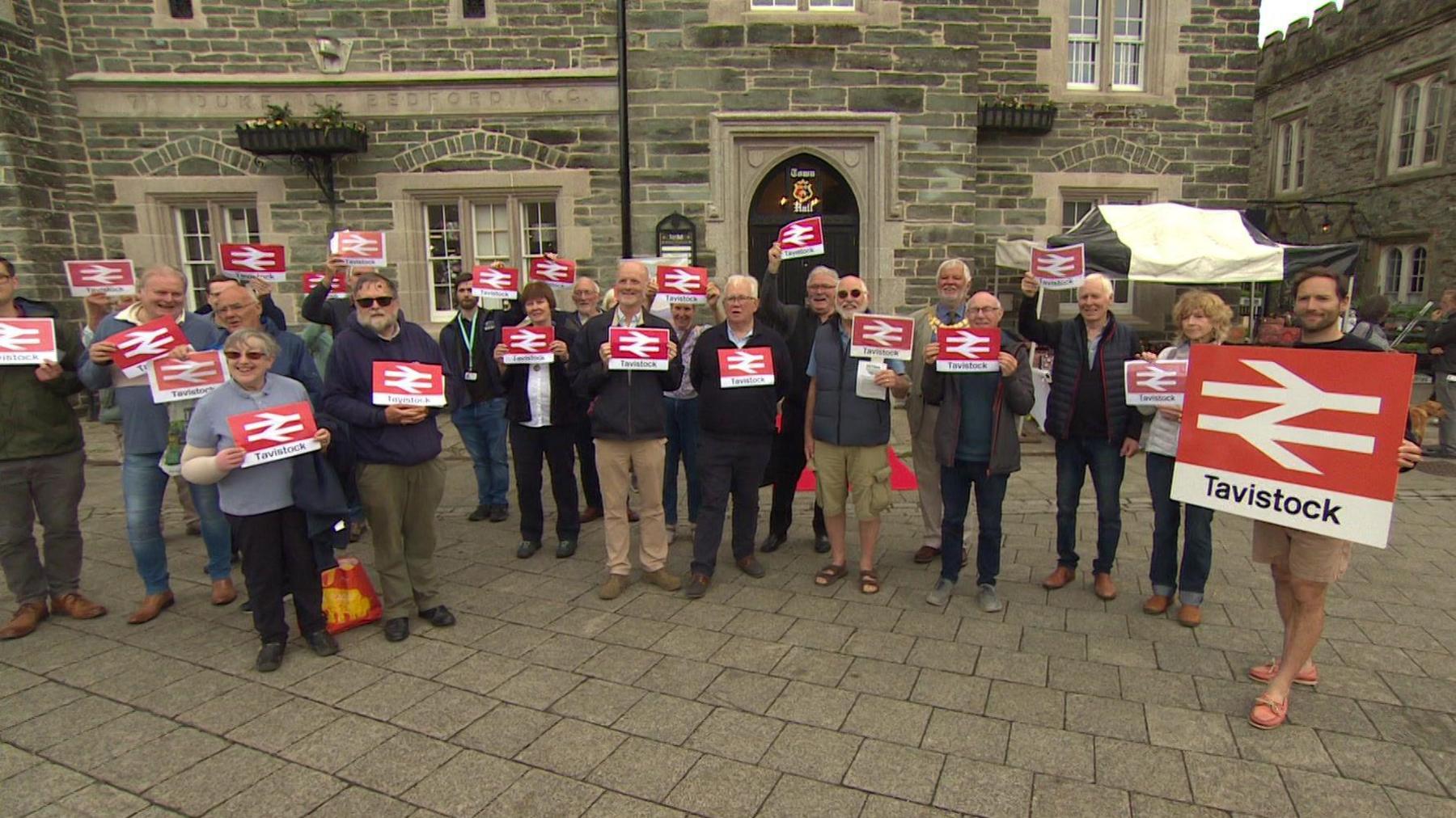 Group of campaigners holding up rail signs saying Tavistock, showing support for the reinstatement of the Tavistock to Plymouth Rail link.