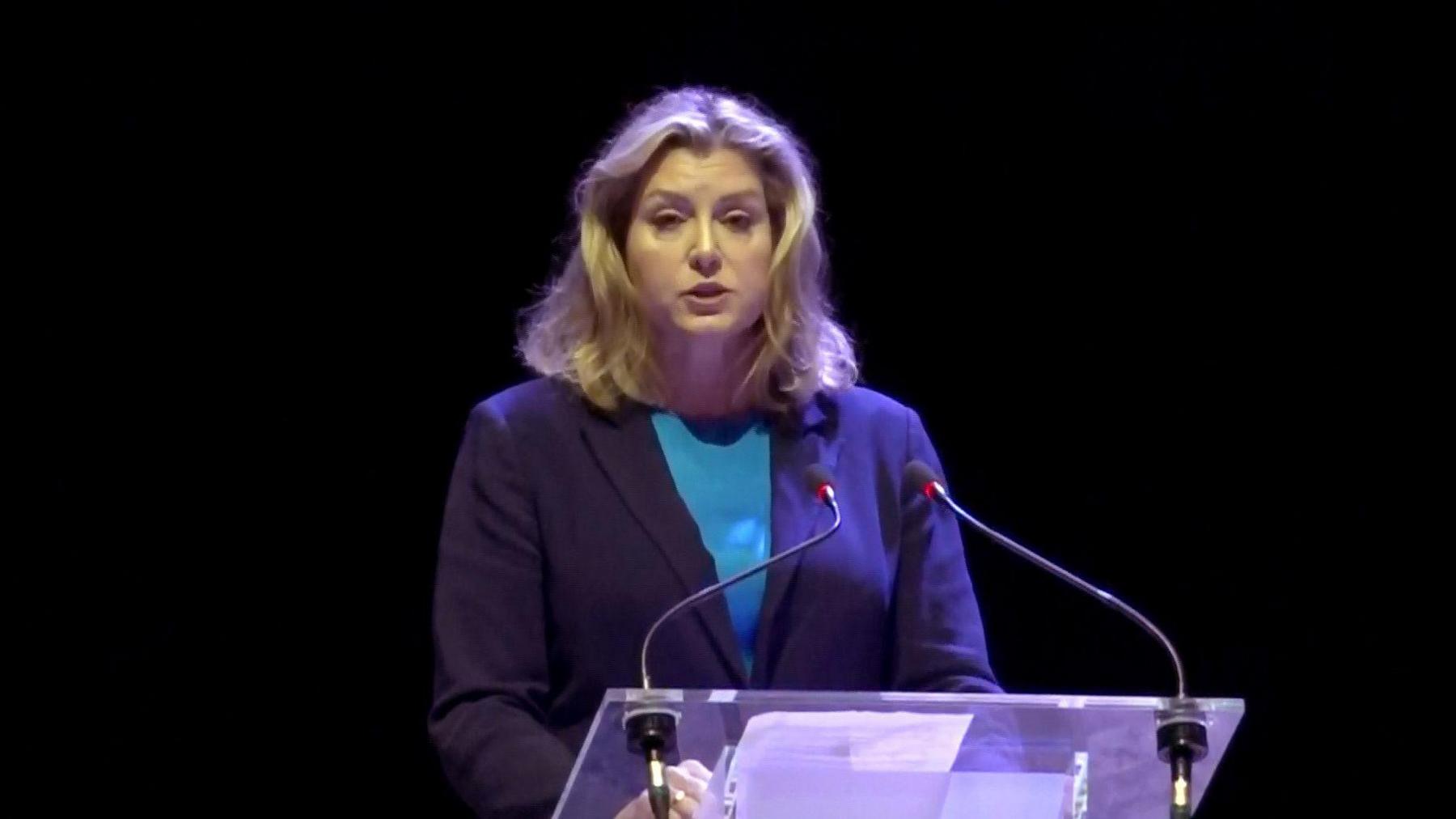 Penny Mordaunt stands at a presentation stand with two microphones. She has mid length blonde hair and wears a dark blue blazer with a lighter blue top underneath.