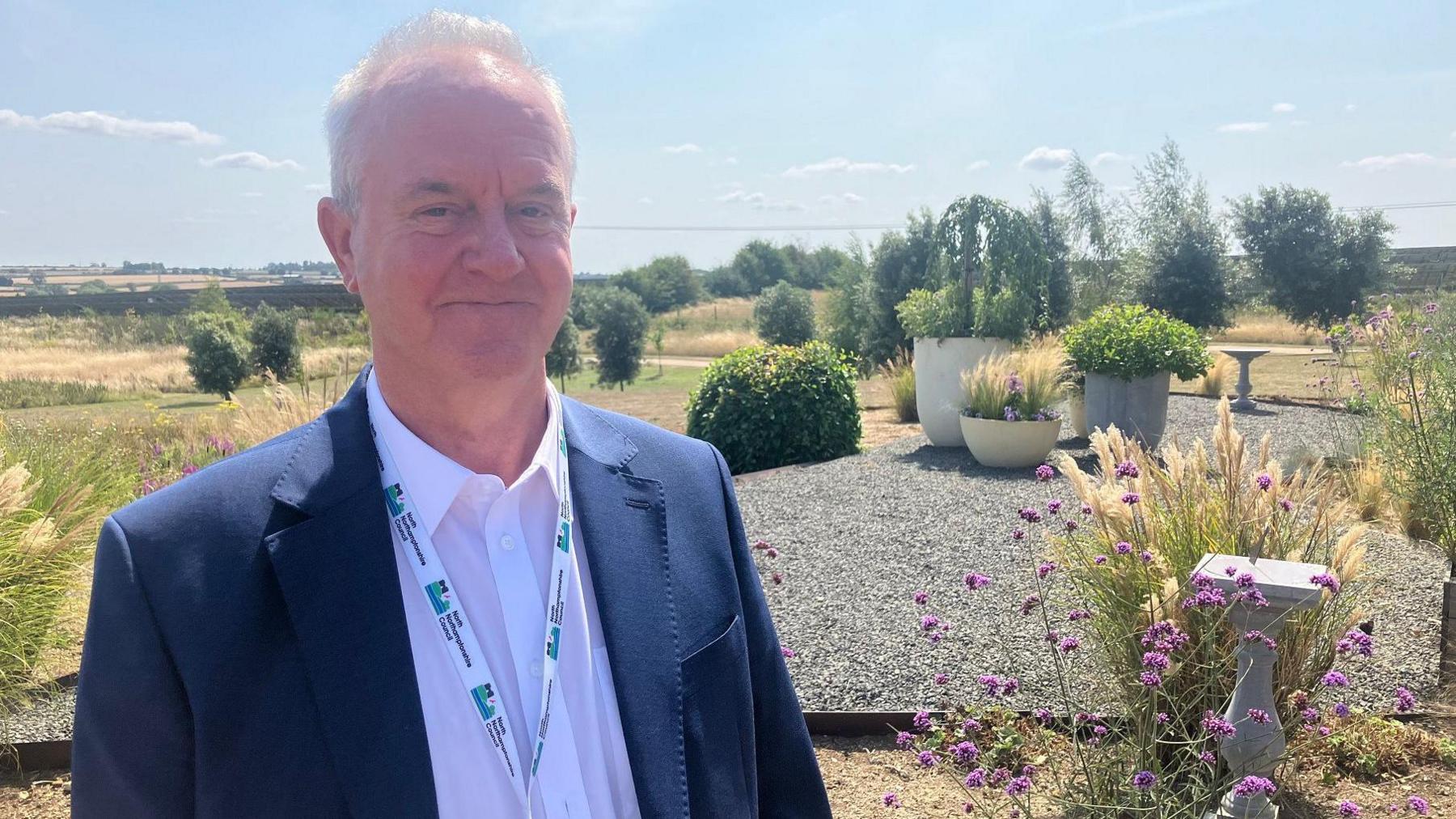 Martin Griffiths smiles at the camera, standing in the crematorium gardens. Behind him are purple flowers and various green shrubs, grasses and bushes as well as distinct gravel areas and garden statues. In the distance are fields and trees. 