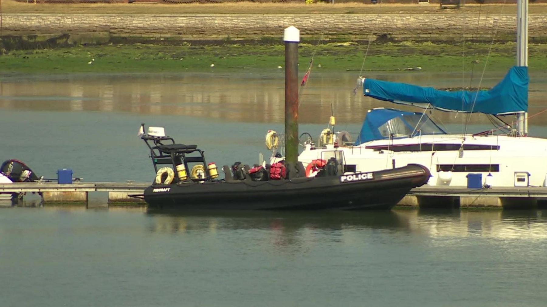 Black police boat a large, black rigid hulled inflatable boat on water alongside a pontoon with other boats in the distance.