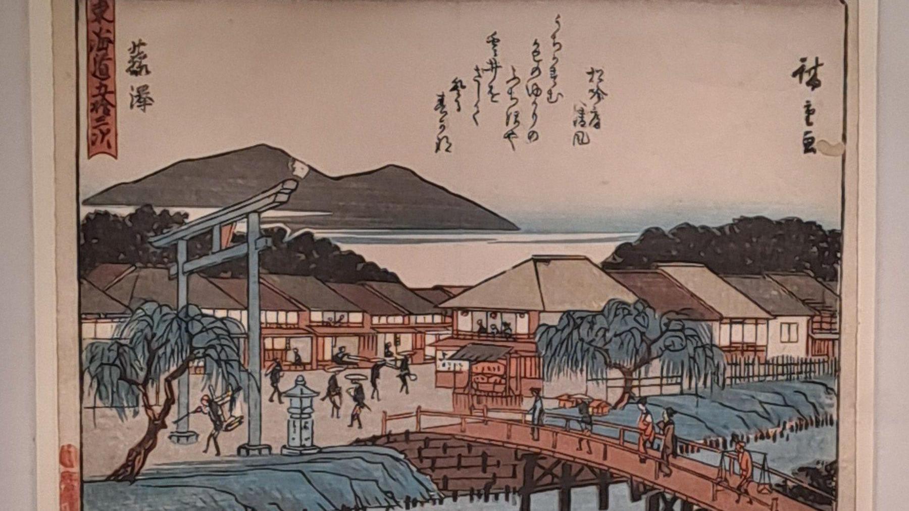 A traditional Japanese woodblock print depicting a scenic town near water. In the foreground, a curved wooden bridge spans a river, with several people walking across it. To the left, a large torii gate and a stone lantern stand near willow trees. 