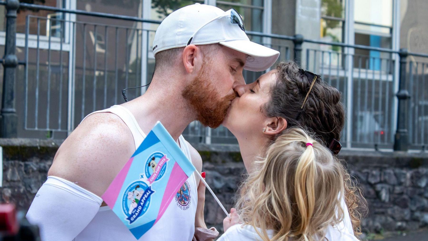 A man and a woman kiss, the woman is holding a young blonde girl in her arms