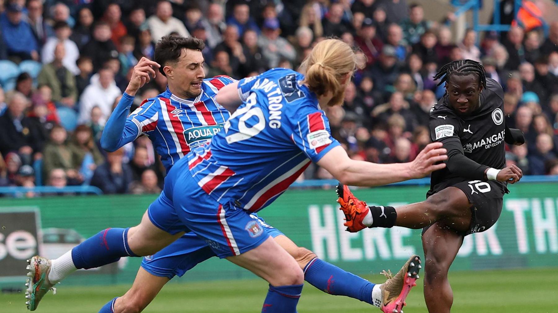 Abdul Abdulmalik shoots with his left foot as two Carlisle players Ryan Galvin and Luke Armstrong - (ARMSTRONG 29) try to block him