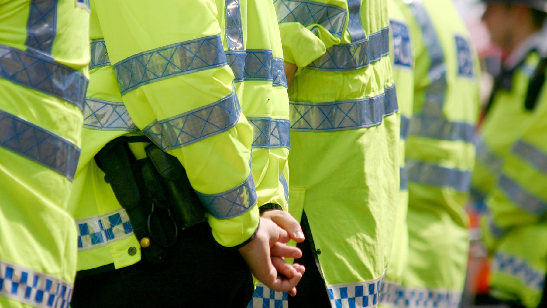 A row of policemen stood outside. Focus is on hands and body of left-most policeman.
