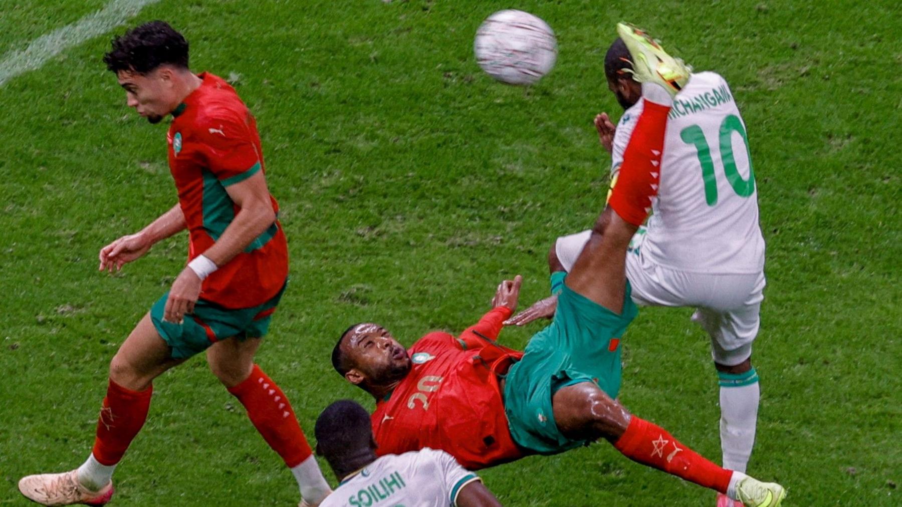 Host Morocco beat stubborn Comoros in AFCON opener Ayoub El Kaabi, wearing red Morocco kit with green trim, connects with a football using his left leg which is raised vertically in the air above him as he flies backwards in the middle of executing an overhead kick. A Morocco team-mate and Comoros defender in white surround him