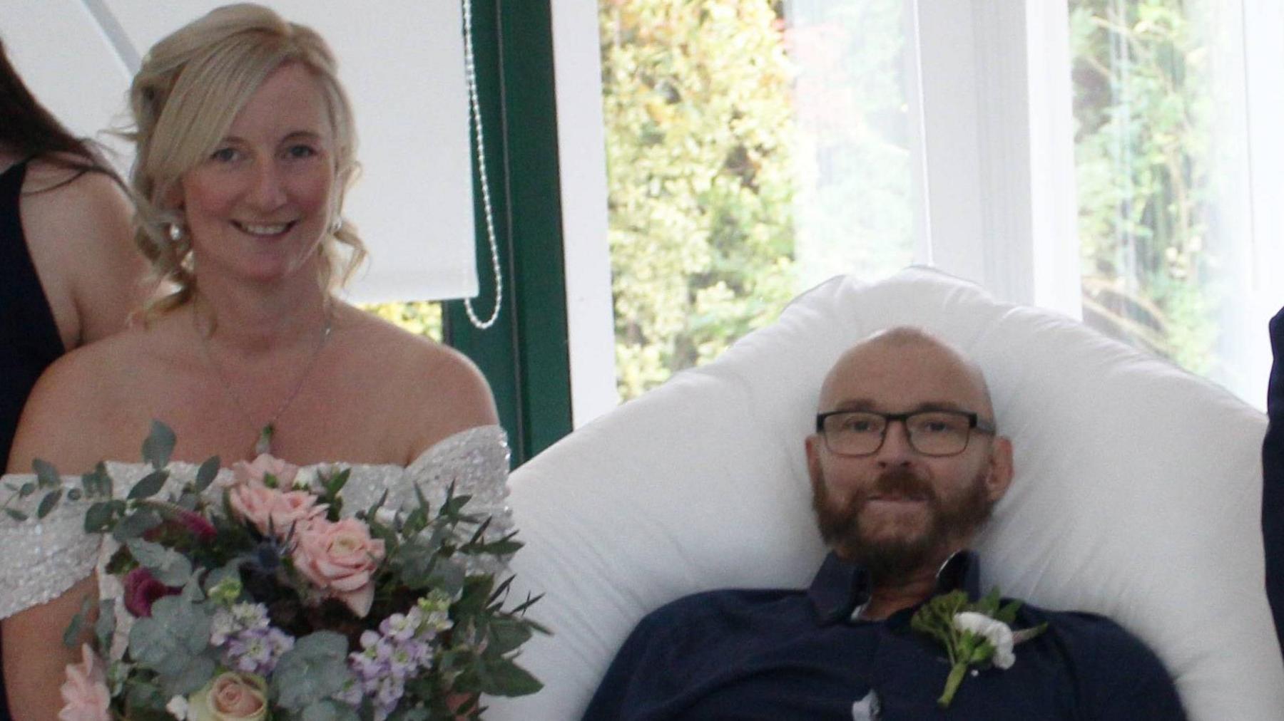 A blonde woman in an off-the-shoulder wedding dress holding a bunch of pink and cream roses and purple flowers, she is standing next to a man in a black shirt propped on a pillow on a bed. He has a cream flower in his lapel. He is wearing glasses and has a dark beard. The pair are both looking at the camera smiling