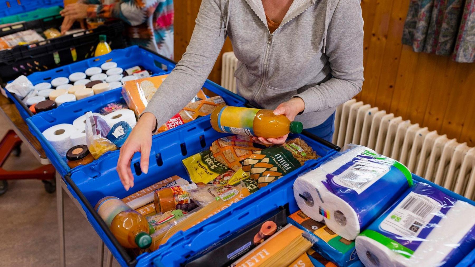 A woman stands over blue trays containing food and household goods such as toilet rolls