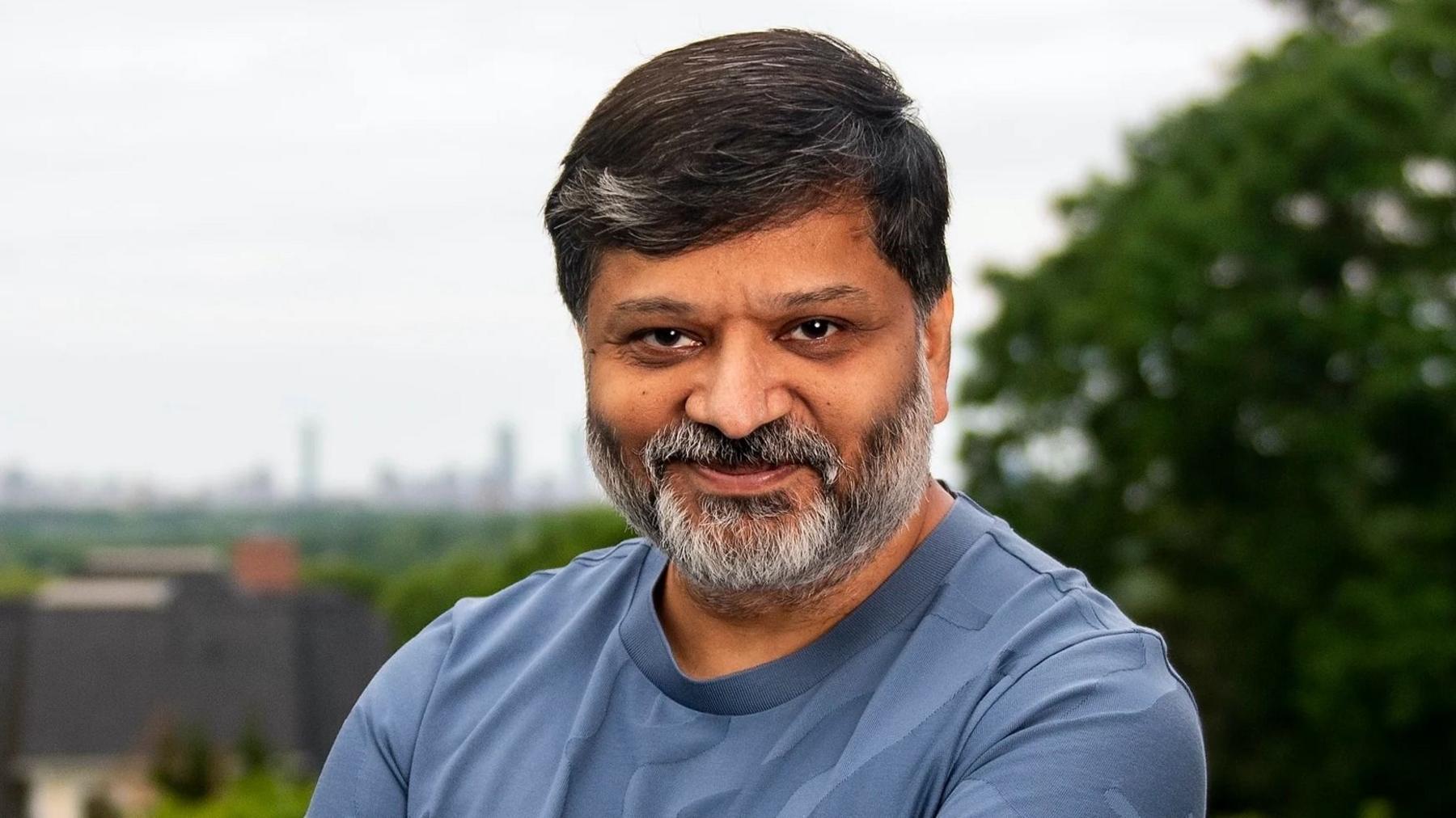 A picture of Dharmesh Shah stood at a slight angle smiling at the camera. He's Indian, in his 50's and wearing a blue t-shirt with black hair and a grey beard.