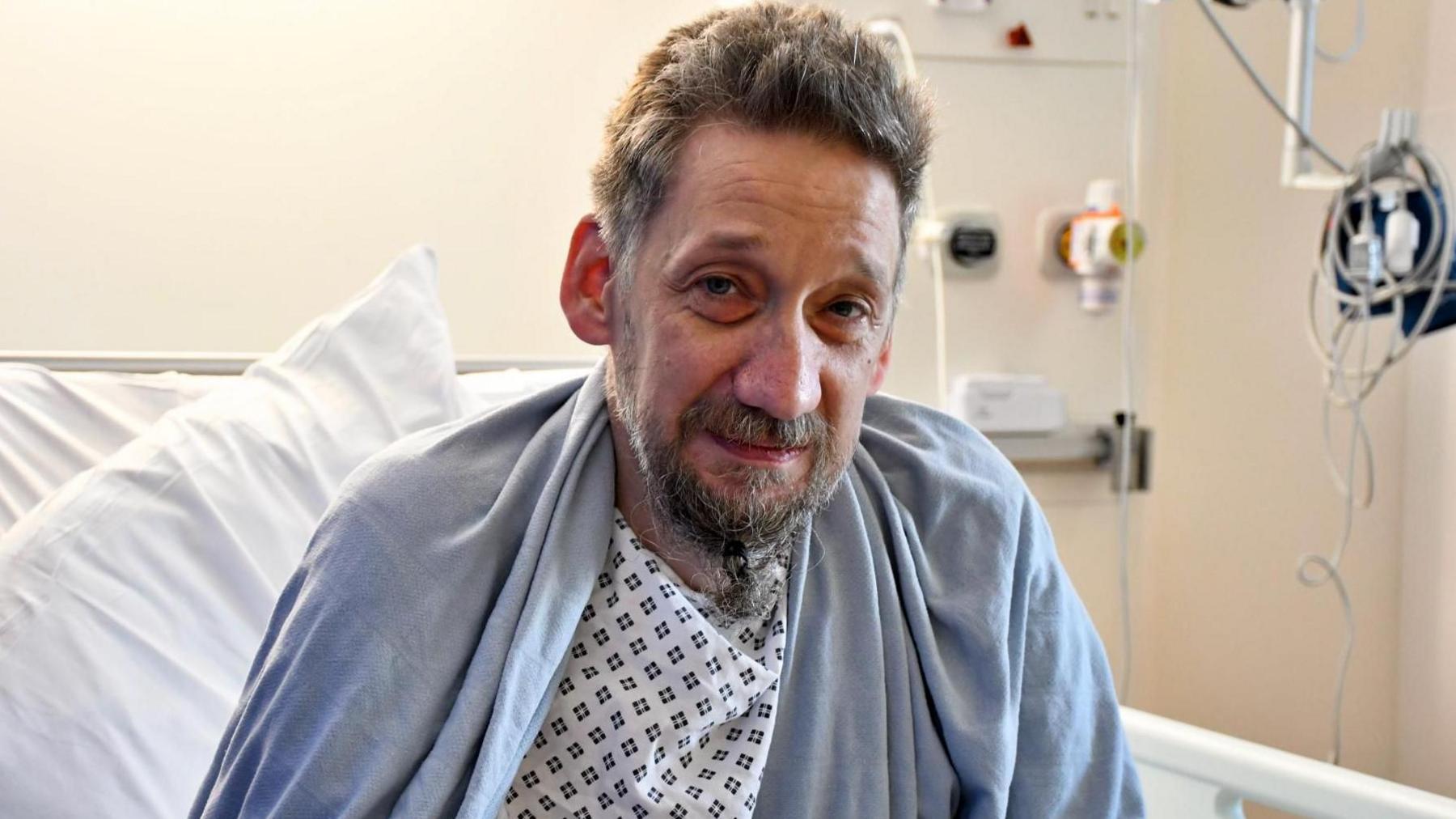 Daniel Evans-Smith looks at the camera from his hospital bed. He is dressed in a hospital gown with a blanket over his shoulders. He had a beard and greying hair. You can see oxygen pipes in the background which are not attached to Mr Evans-Smith