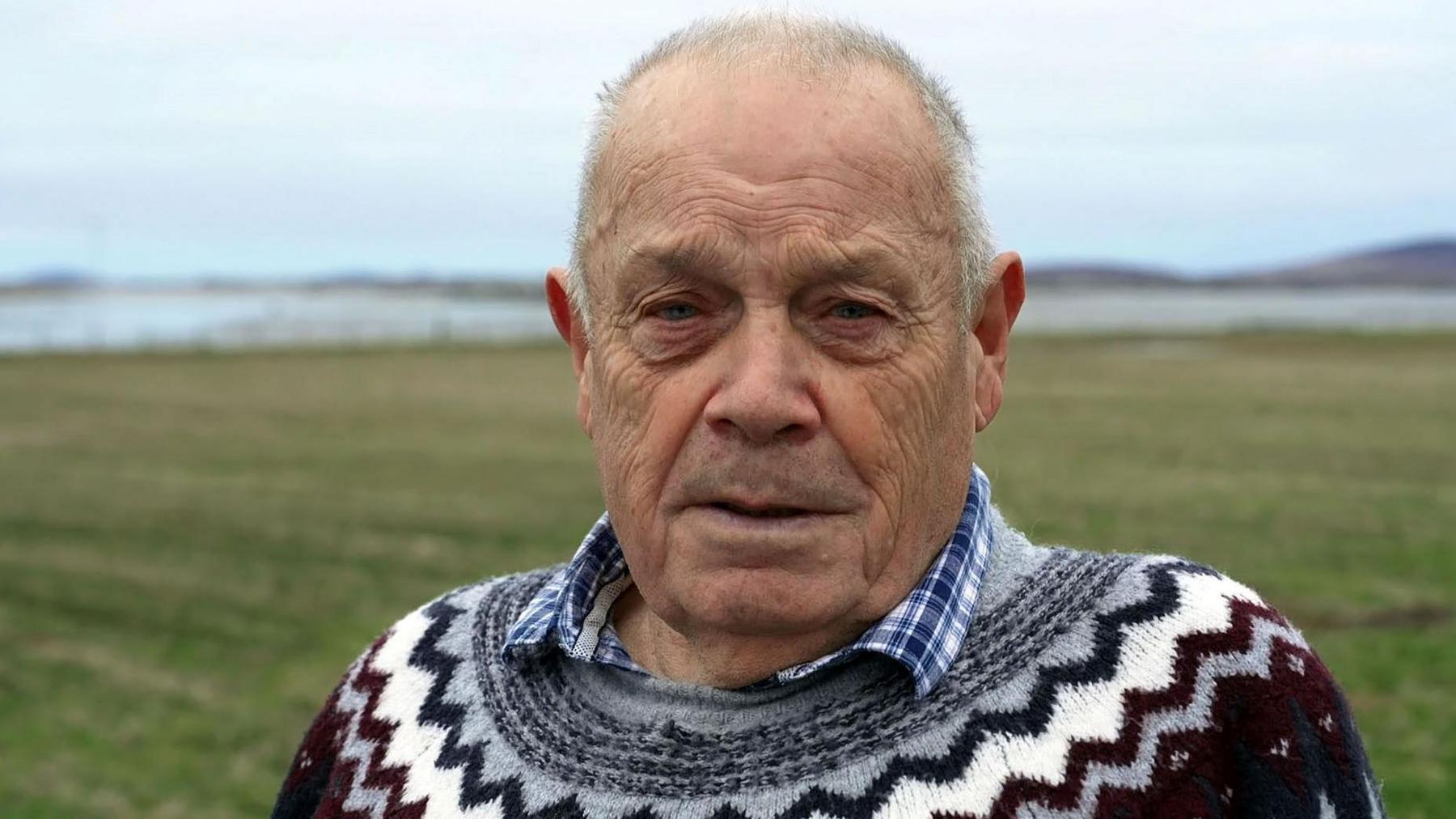 David is looking into the camera. He is wearing a patterned jumper. He is standing in an area of coastal meadow called machair.