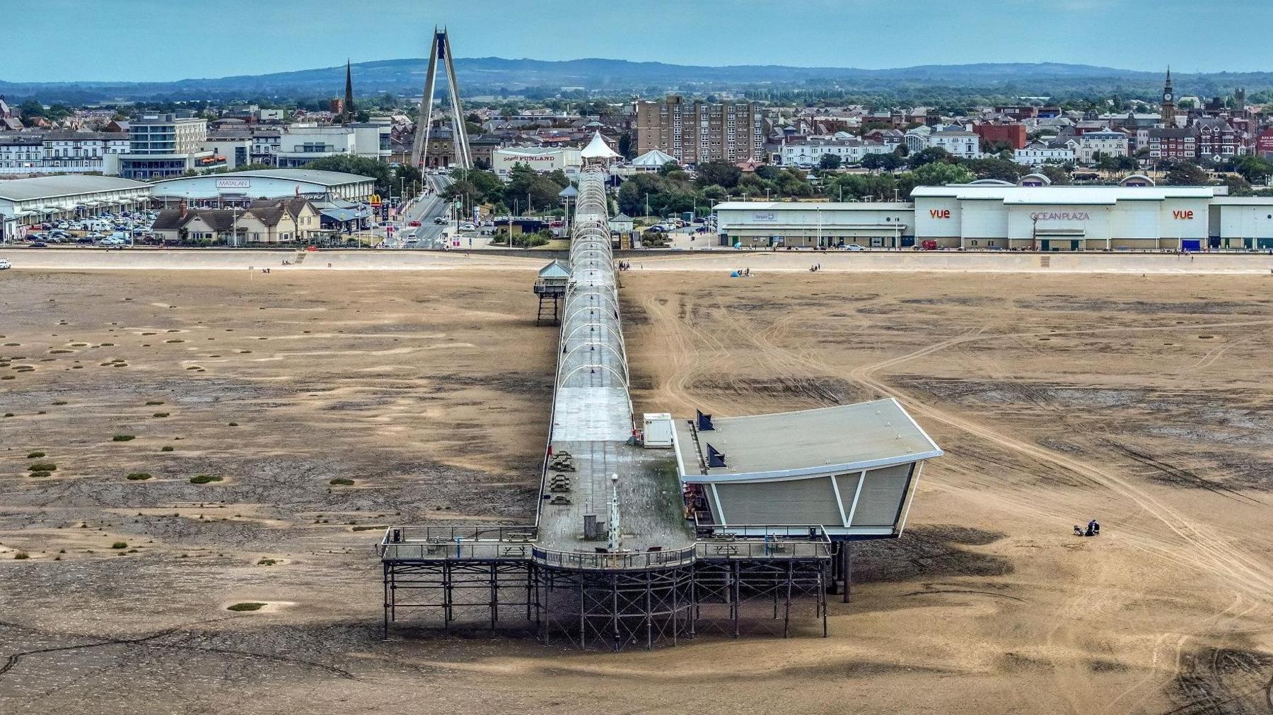 Long empty pier stretches over the sandy beach down the centre of the image. Shops, leisure venues, a tall fairground ride can be seen in the background along with hills rising in the backdrop.