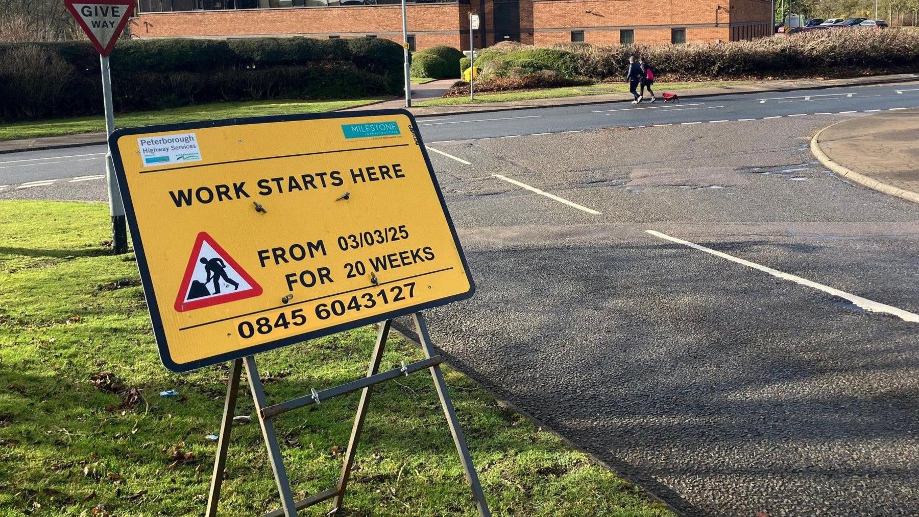 A yellow roadworks sign reads 'Work starts here from 03/03/25 for 20 weeks'. It is situated on the grass next to a junction at Thorpe Wood. The signage also has logos for Peterborough Highways Services and Milestone Infrastructure Limited, who will deliver the works.