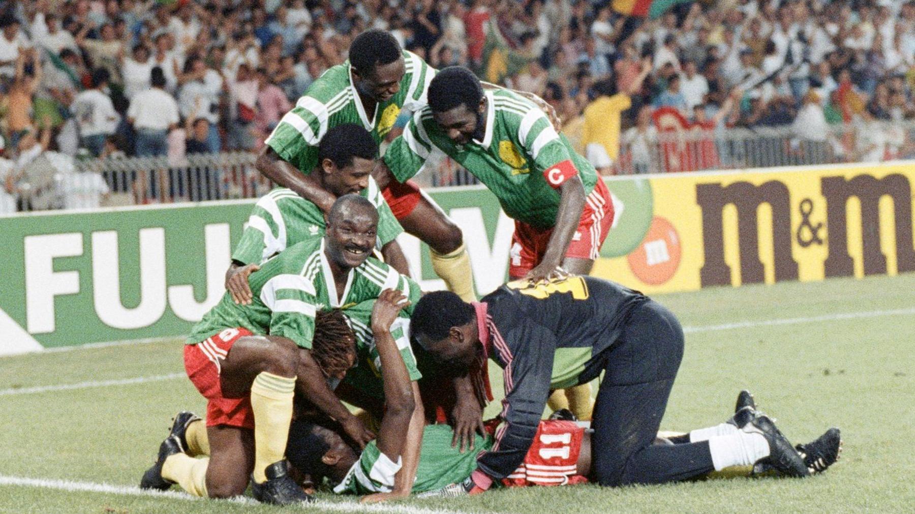 Cameroon outfield players in green shirts, red shorts and yellow socks, as well as the goalkeeper dressed mostly in black including his tracksuit bottoms, celebrate a goal at the 1990 World Cup by piling on top of Eugene Ekeke, who lies flat on his back as six players gather around him and offer congratulations. Advertising hoardings can be seen in the background, with a packed stand of fans out of focus in the distances. One player, Roger Milla, looks up from the pack and smiles towards the camera
