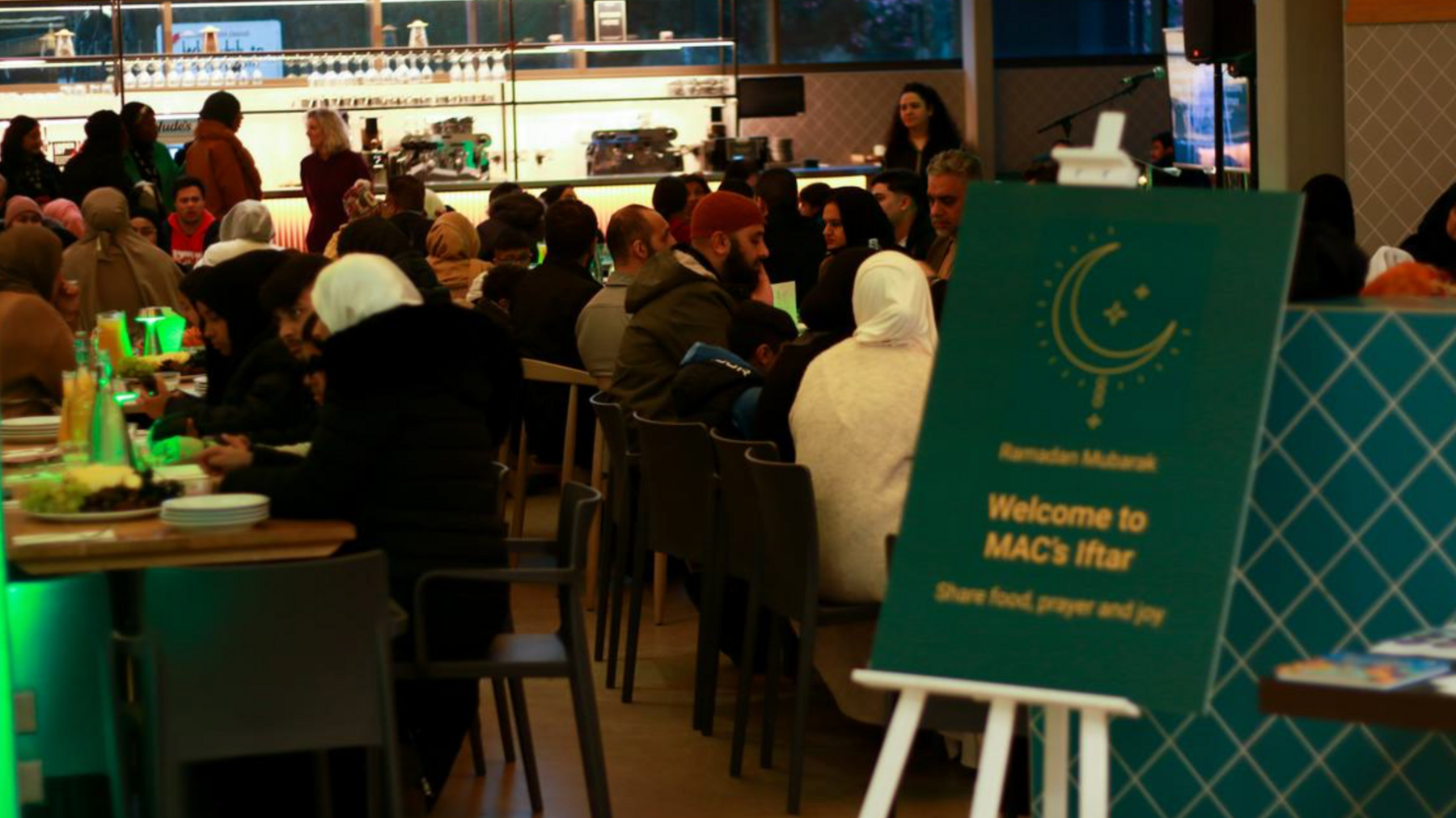 Rows of people wearing religious clothing sat down and eating. A green sign says Welcome to Mac's Iftar.