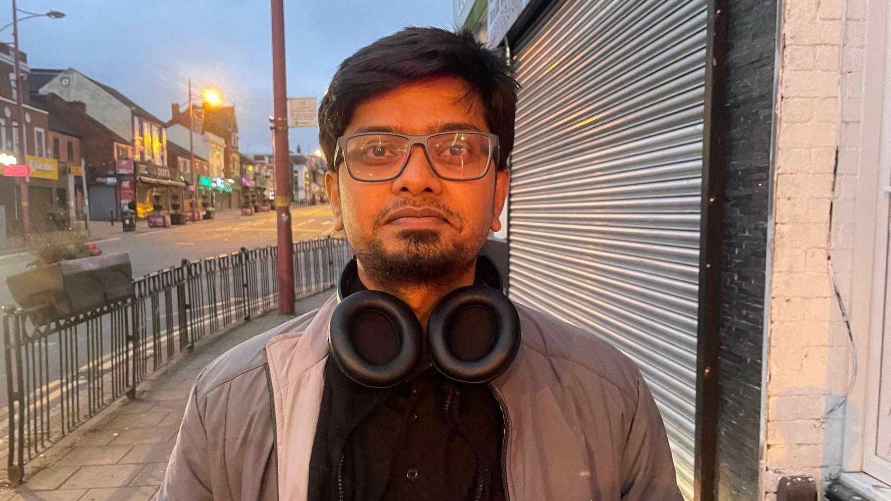 Mariaj Khan stands in the Soho Road in Handsworth. He is wearing a black top and beige jacket and has headphones around his neck. He has glasses and a beard. There are shops in the road behind him and there is a planter on railings by the street coroner. It's early and just getting light.