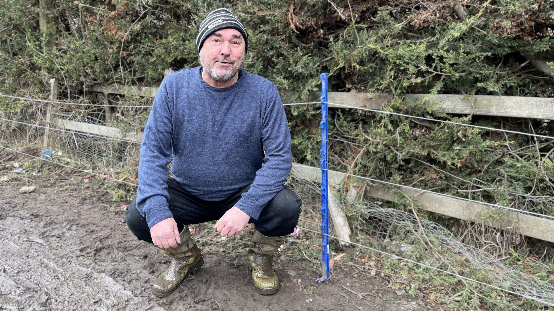 Farmer Trevor Armiger crouched by an electric fence which has thin blue pylons. He is wearing wellington boots, a blue jumper and a stripy hat. He has a short grey beard