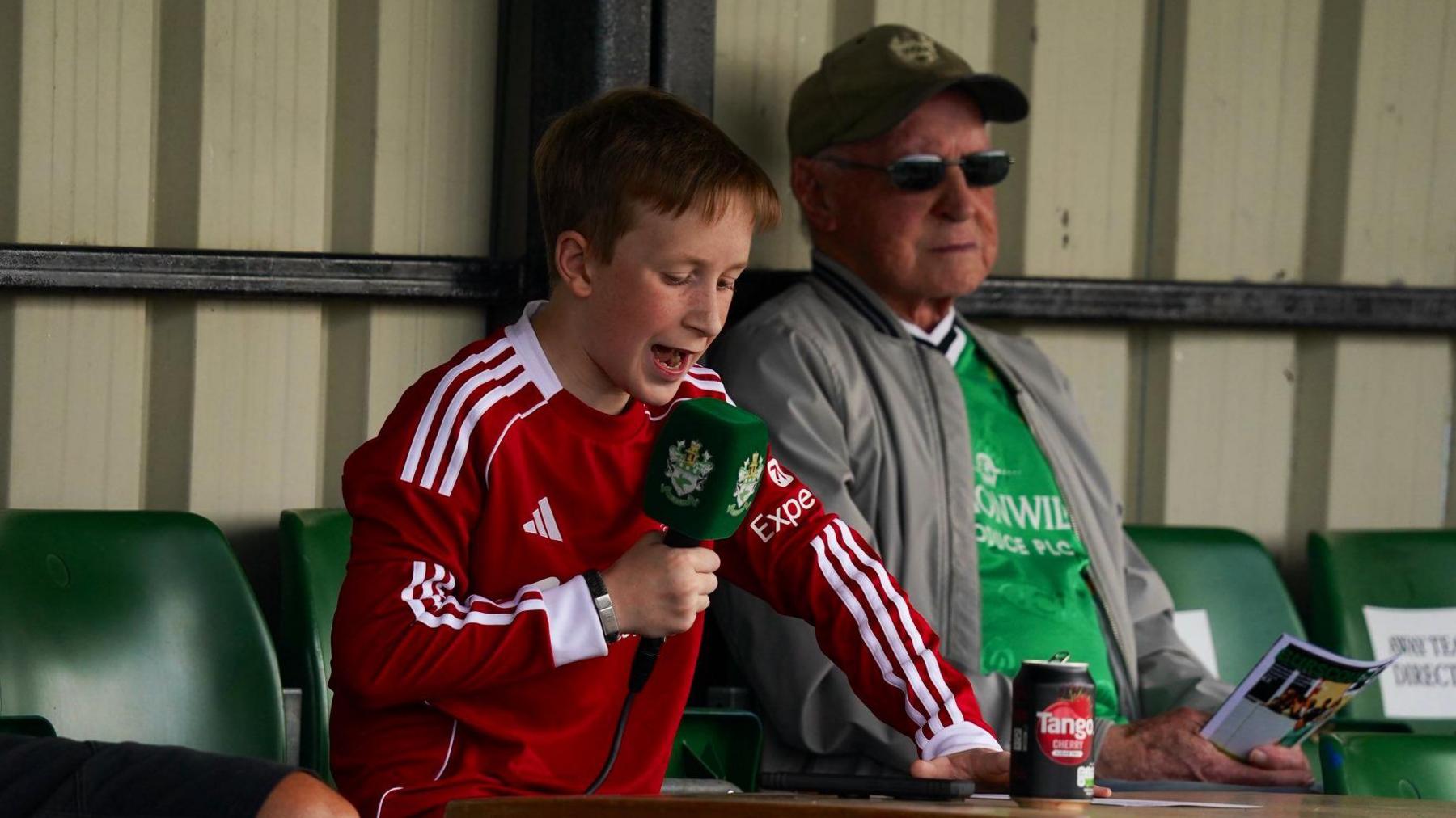 James leans forward as he announces names from a sheet on a desk which a fizzy drink can on it. He is holding the microphone and standing next to an elderly man wearing the green Burscough FC shirt under a grey jacket. They are at the back of a stand with rows of green seats.